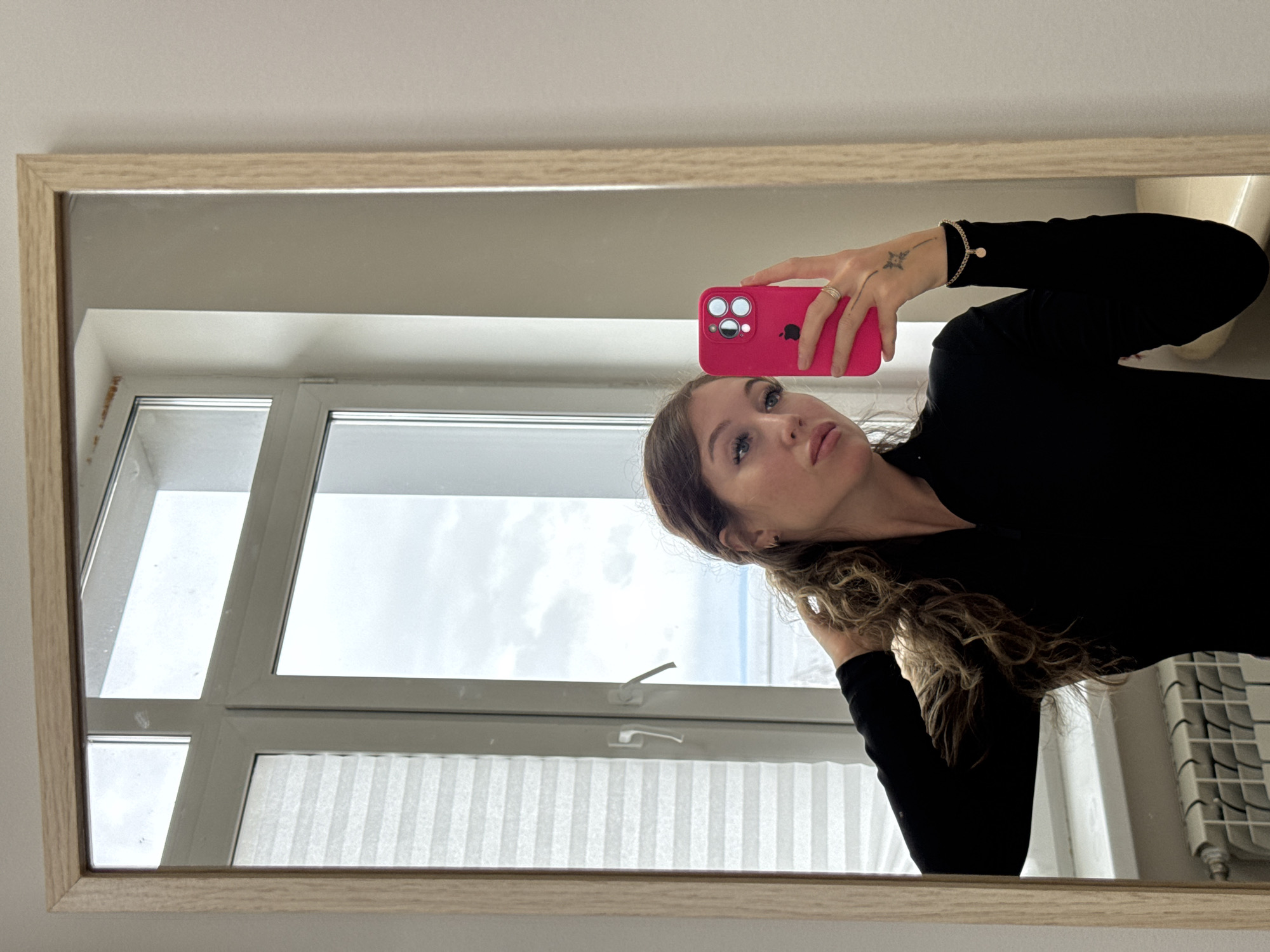 The original image is oriented 90 degrees counter-clockwise. When viewed upright, it depicts a close-up mirror selfie of a woman indoors.
The primary subject is a woman, visible from the chest up, with long, wavy brown hair. She is looking slightly upward and to the viewer's left, with a neutral and posed expression. Her eyes are light-colored, and her eyelashes appear prominent. She wears a black long-sleeved top. Her right hand, adorned with a ring on her ring finger and a small, star-like tattoo on the back near her thumb, holds a bright pink smartphone with a matching case, capturing her reflection. A delicate bracelet with a small round charm is visible on her right wrist. Her left hand is raised towards her hair, appearing to adjust or pose with it.
The setting is an indoor space reflected in a large mirror with a light wooden frame. To the left of the woman, through the mirror, a large window is visible, featuring white frames and a handle, suggesting it can open. The sky outside the window appears overcast and bright. Below the window, part of a white pleated blind is visible. To the right side of the woman, also reflected in the mirror, a portion of a white radiator with a grid-like pattern can be seen.
The activity is a self-portrait taken using a smartphone's front-facing camera, reflected in a mirror. The lighting is bright, consistent with daytime, likely coming from the large window. The only visible text is the partially obscured Apple logo on the back of the pink phone case.