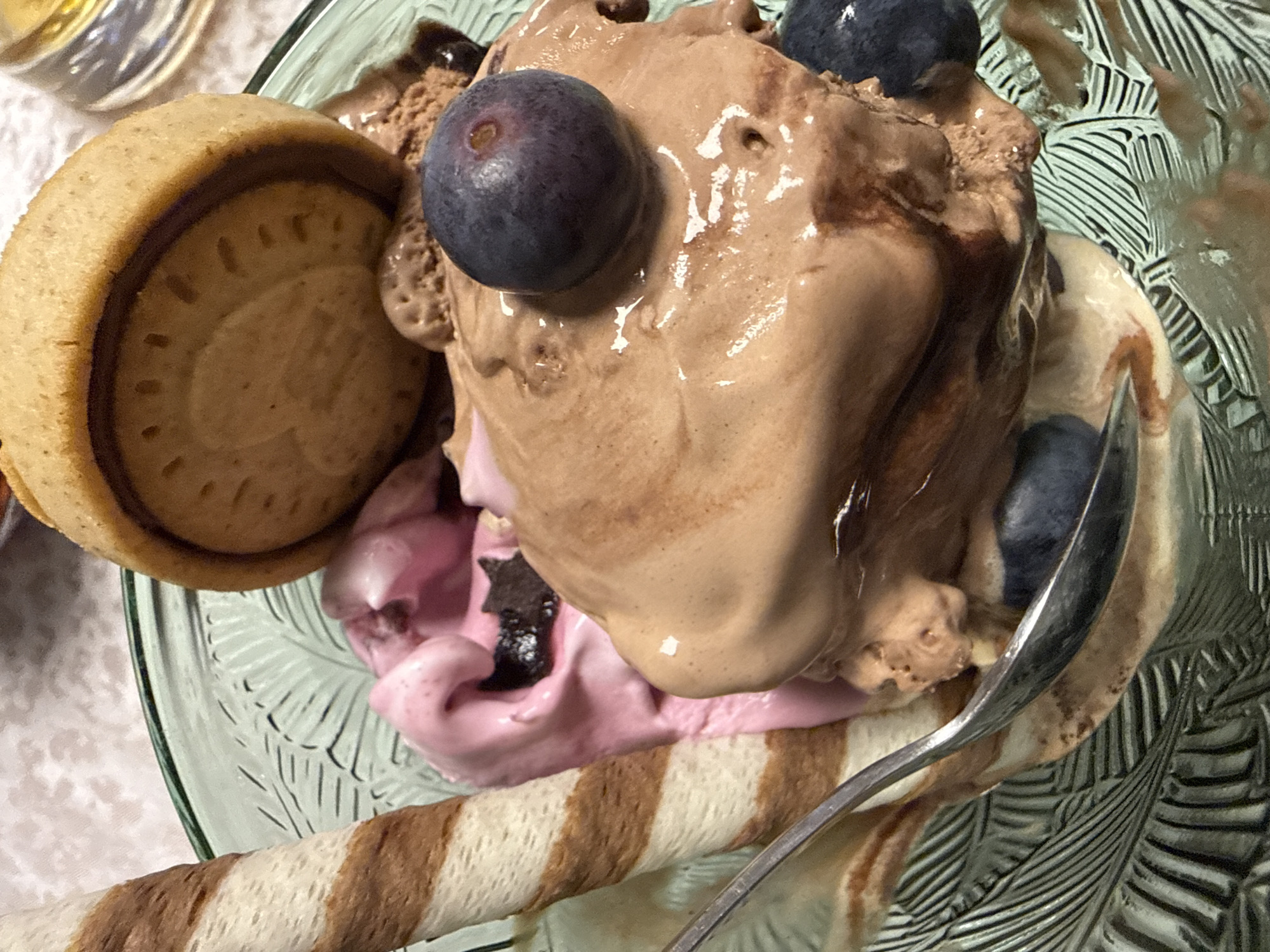 A close-up overhead shot shows an ice cream sundae in a green glass dish. The sundae features scoops of chocolate and strawberry ice cream, garnished with two blueberries atop the chocolate scoop. A chocolate-filled cookie rests against the side of the ice cream. A striped wafer stick lies along the bottom of the dish, and a spoon is placed to the right of the ice cream. The background consists of a white surface and hints of what appears to be another glass object. The location of the photo is unknown, and no details connect the subject to Varna, Bulgaria.
