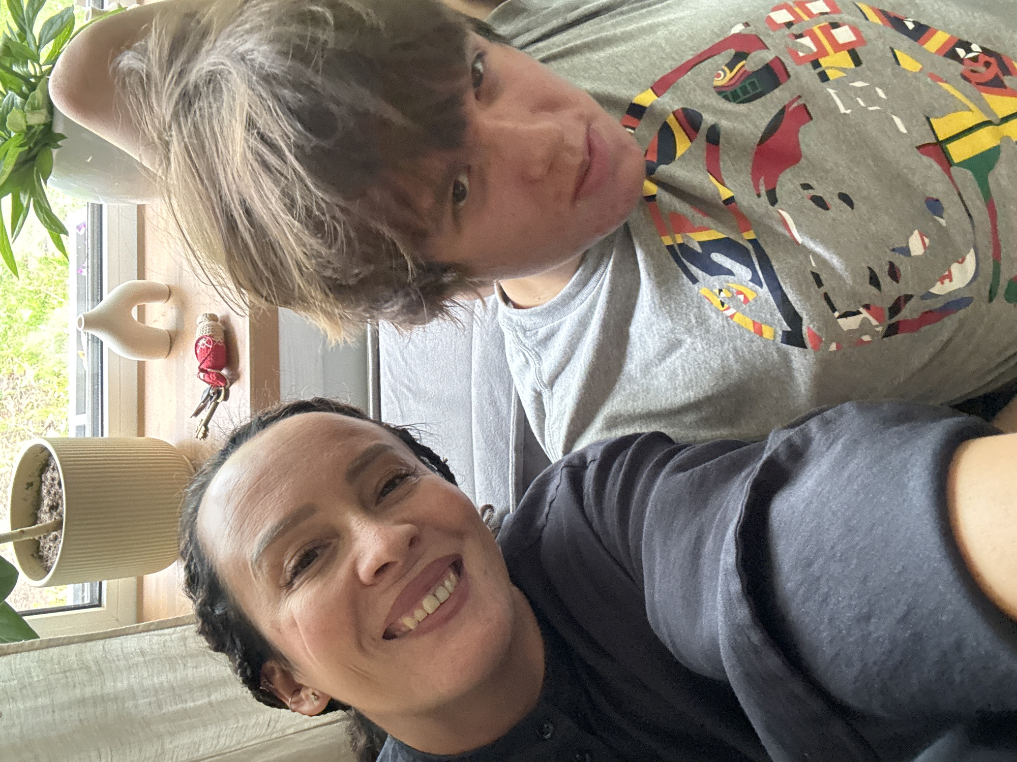 This casual, indoor selfie, captured in Chaikovsky, Russia, features two individuals in a relaxed home setting, illuminated by natural daylight. The image is oriented sideways, suggesting it was taken with the camera rotated.
In the foreground, an adult female with medium skin tone and dark hair, possibly styled in braids or dreadlocks, smiles warmly, directly at the camera, revealing her teeth. She wears a dark gray or black short-sleeved top.
Behind and slightly above her, a younger individual with light brown, shaggy hair and dark eyes gazes towards the camera with a neutral or subtly curious expression. They are dressed in a light gray short-sleeved t-shirt adorned with a vibrant, abstract, and geometric pattern featuring shapes in red, yellow, black, blue, and white.
The background elements suggest a domestic space. To the left, a window provides ample natural light, showing green foliage outside, confirming it is daytime. Below the window, a light-colored curtain is visible. On a wooden surface, likely a windowsill or shelf, a white ribbed plant pot holds a green plant. Nearby, a small, white, U-shaped decorative object is placed, and a set of silver keys is attached to a small, red, crocheted or knitted pouch. To the right, behind the foreground subject, a gray, textured fabric, possibly upholstery from a couch or bed, is partially visible.
There is no visible text in the image.