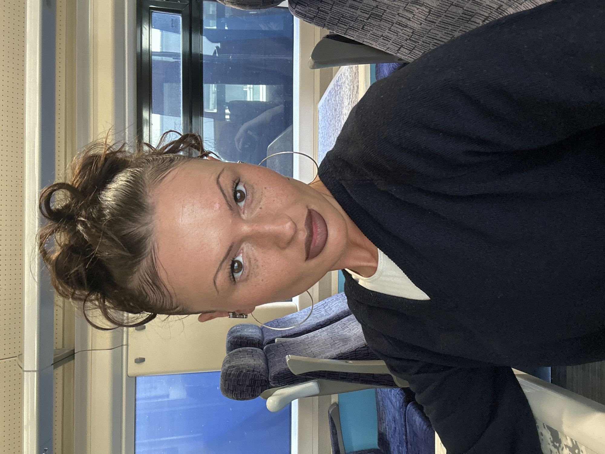 A medium shot depicts a woman on a train, likely in London, United Kingdom. She has brown hair in an updo, wears large hoop earrings, and is dressed in a dark blue sweater over a white top. The train carriage has blue and gray patterned seats and large windows showing a blurred urban landscape. The lighting is diffused, suggesting daylight.