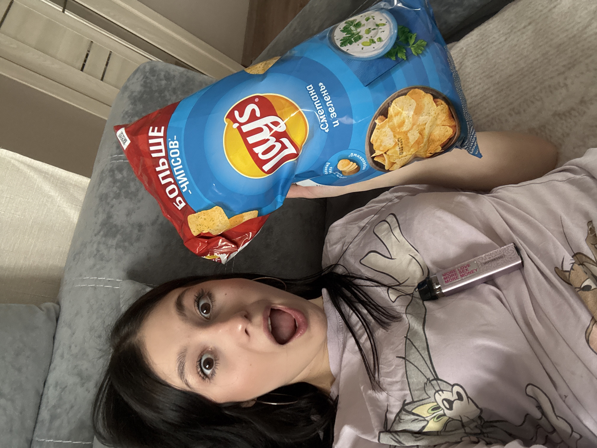 A young female with dark hair and hoop earrings is depicted in an indoor setting, identified as Chaikovsky, Russia. She is lying on a grey upholstered couch, positioned with her head towards the top left of the frame. She wears a light-colored t-shirt featuring a cartoon print of "Tom and Jerry." Her eyes are wide, and her mouth is open, indicating a surprised expression. Her right arm is visible, holding a large, unopened bag of Lay's potato chips. The chip bag is predominantly blue and red, displaying the Lay's logo and the flavor "Сметана и зелень" (Sour Cream and Greens) in Cyrillic. The phrase "БОЛЬШЕ ЧИПСОВ" (MORE CHIPS) is printed in red and white at the top of the bag. A silver-colored Vaporesso vape device, inscribed with the text "MORE LOVE MORE SEX MORE MONEY," rests on her shirt near her chest. The background shows a light-colored wall and light-toned curtains. No other individuals or interactions are observed.