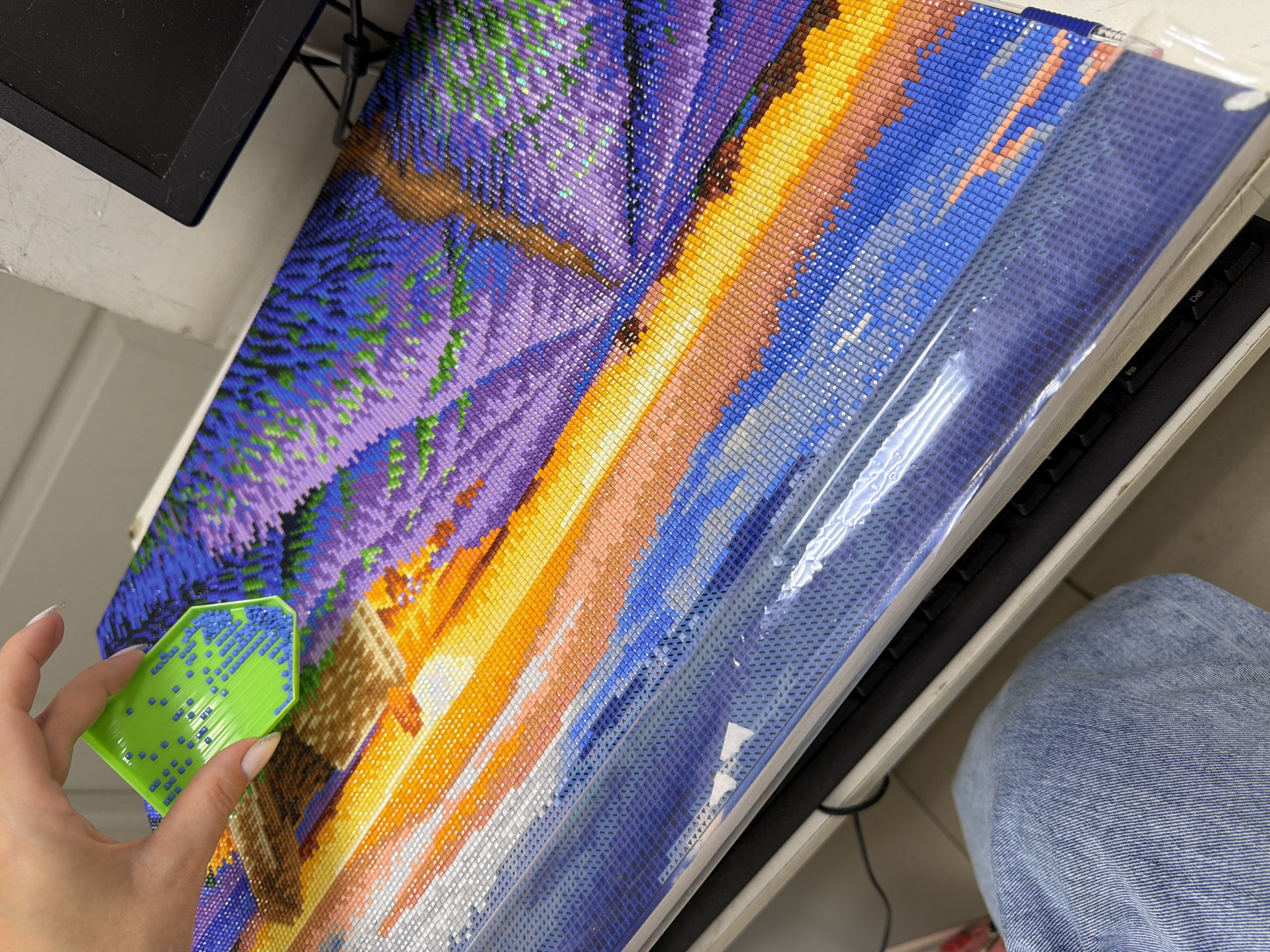 A person holds a green tray filled with blue rhinestones over a partially completed diamond painting depicting a landscape scene with purple and blue hues, suggestive of a sunset or lavender field. The painting is covered with a clear protective film in sections. A portion of a black computer monitor and keyboard are visible at the edge of the frame. The person is wearing denim jeans.
There is no evidence to suggest the location is in Chaikovsky, Russia.
