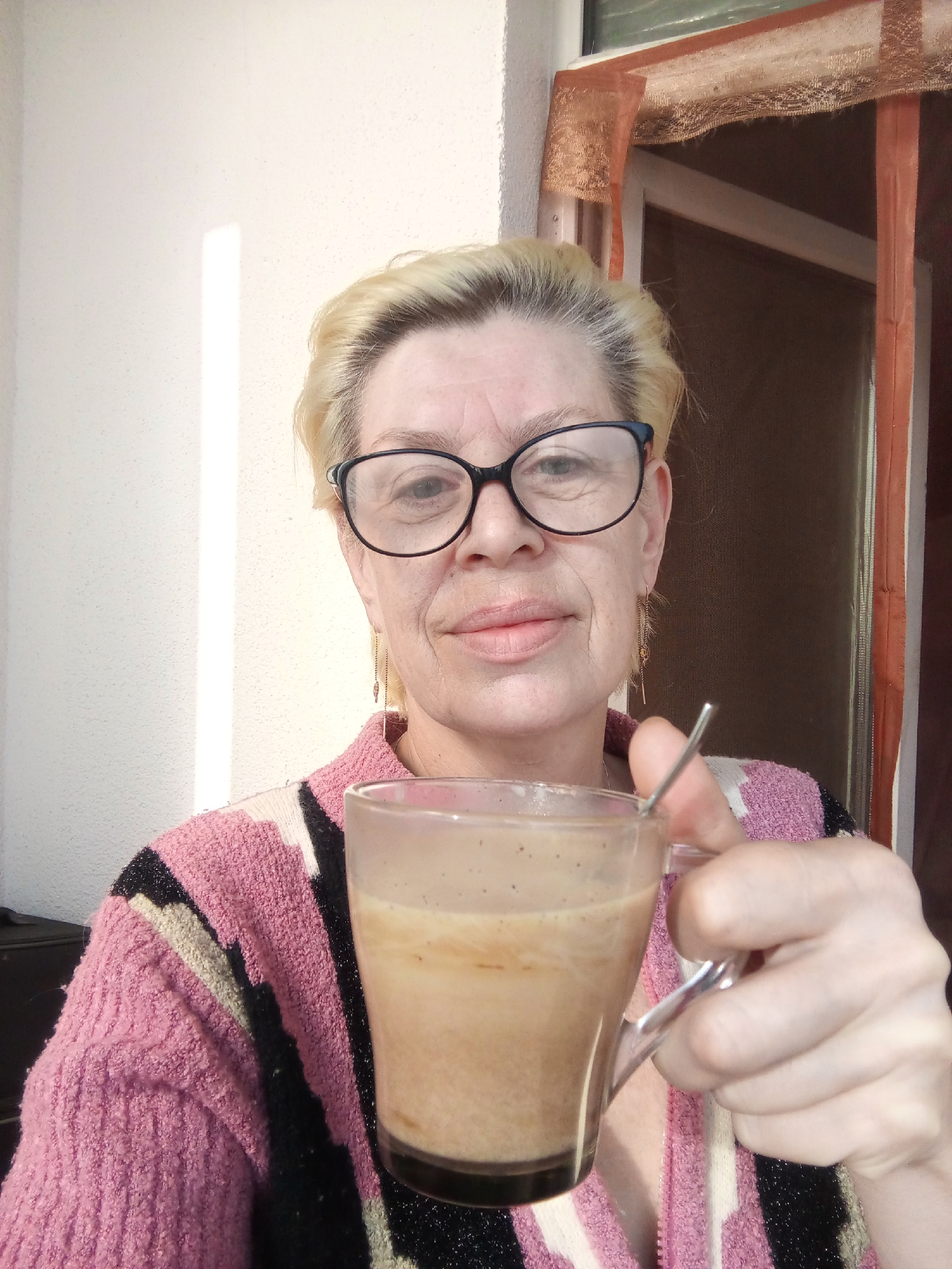 A blonde, fair-skinned woman wearing black-framed glasses, a pink, black, and white sweater, and drop earrings is holding a clear glass mug filled with a beige liquid, possibly coffee or tea, in an indoor or semi-outdoor setting. A small metal spoon is visible within the mug. A sunlit white wall is to the left of the woman, and a window or door with a screen and a light brown curtain or mesh is behind her on the right. The location is specified as the city of Kommunar, Russia.