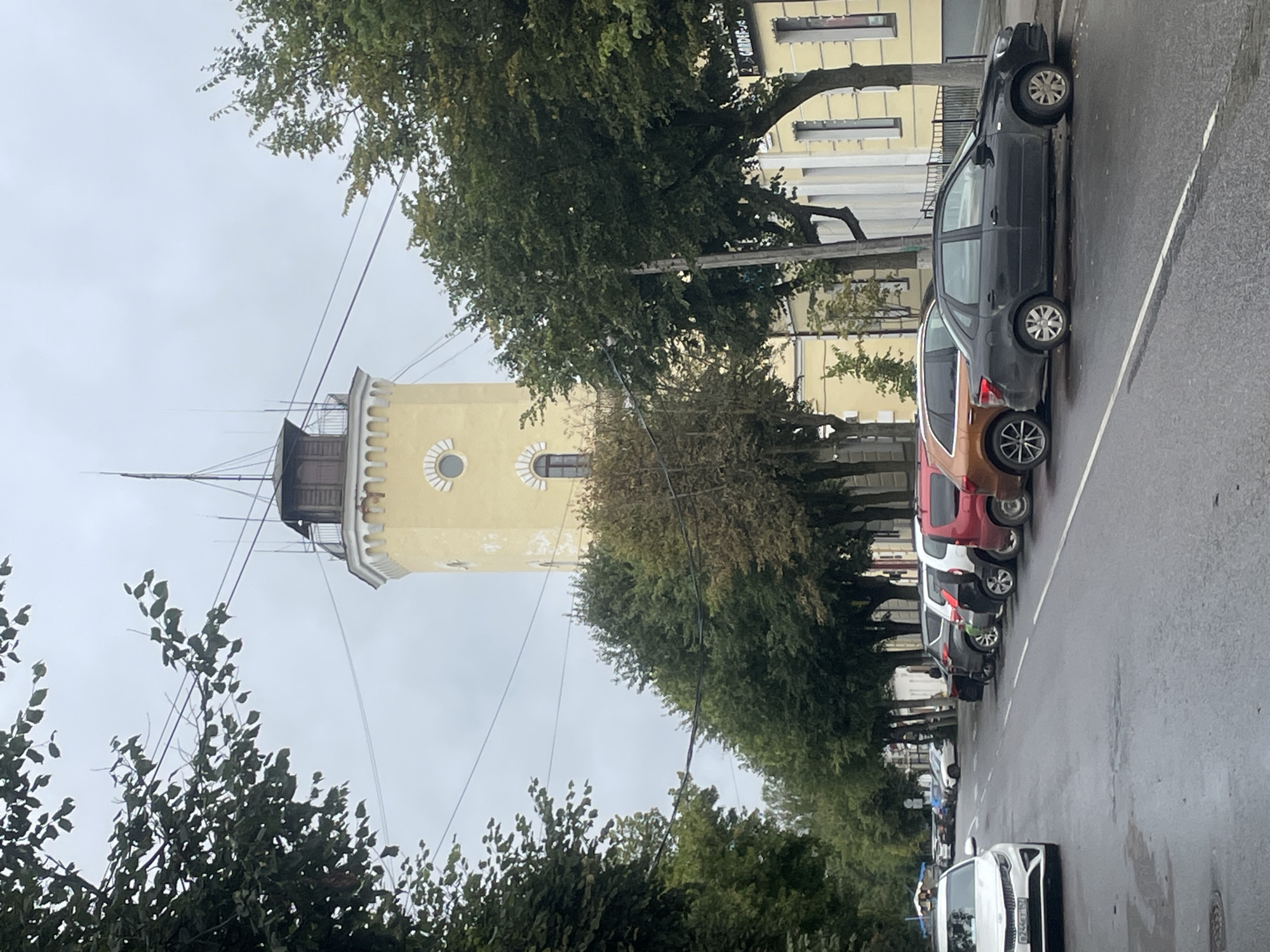 A view from a street in Gatchina, Russia, shows a yellow fire tower in the background, partially obscured by trees. The tower has a dark-capped top with an antenna and a viewing platform. It features arched windows and decorative corbels. The street is lined with cars parked on the right side, and trees on both sides. The road appears wet, suggesting recent rain. Buildings with yellow facades and white trim are visible behind the trees. A white Kia sedan is parked on the left side of the street, facing away from the camera. Several other cars, including a dark sedan and a red SUV, are parked in a row on the right. The sky is overcast.