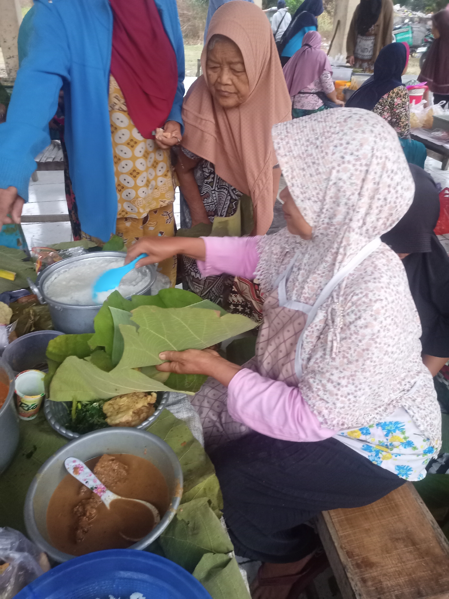 In Nganti, Indonesia, a group of women are gathered around a food stall, preparing and serving dishes. A woman in a pink hijab and patterned shirt uses a blue ladle to scoop white food from a pot. She is seated on a wooden bench. Another woman, wearing a maroon hijab and yellow patterned dress, stands nearby, looking at the food. A third woman, in a peach hijab, also appears to be involved in the preparation. Large green leaves are used as serving platters for various food items, including what looks like rice and a brown curry. The scene suggests a communal meal or a market setting.