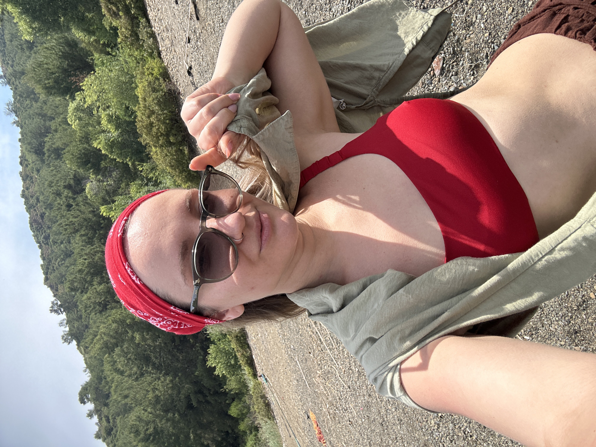 A woman is lying on a rocky surface outdoors, taking a selfie. She is wearing a red bandana, sunglasses, a red bikini top, a tan-colored shirt, and brown shorts. Trees are visible in the background. The photograph appears to be taken in an outdoor setting, but there is insufficient information to establish the location in the city of Fethiye, Turkey.