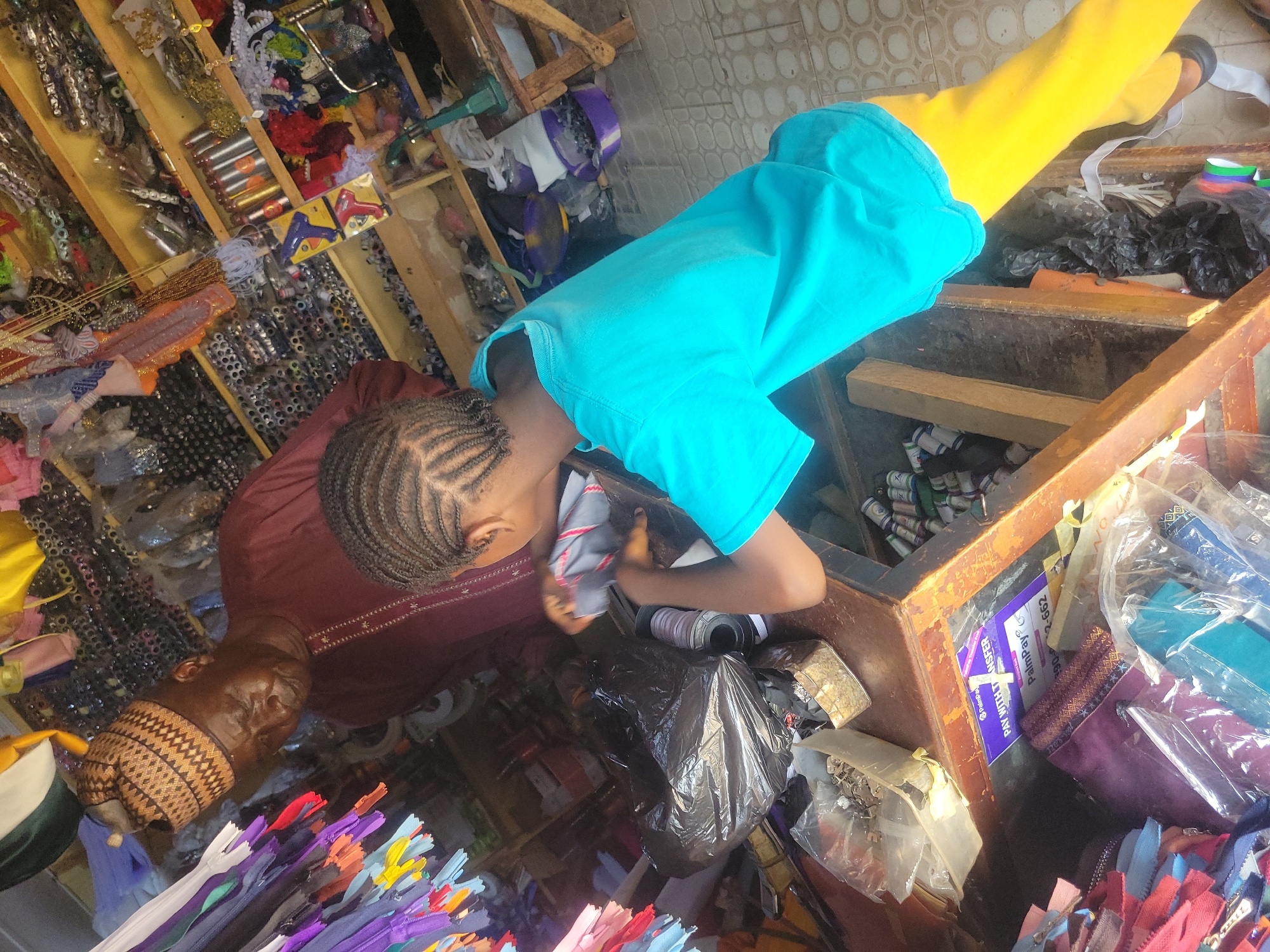 A top-down, canted view shows two individuals in a cluttered store. The location is implied as Jos, Nigeria, though no explicit indicators exist in the image.
A person with cornrow braids and wearing a turquoise t-shirt leans over the edge of a brown, wooden counter. Their hands are inside the counter, possibly interacting with items within. The counter appears to hold a collection of fabric, spools, and possibly other sewing or craft-related materials. A black plastic bag sits near the person's hands.
Next to this person, and closer to the bottom of the frame, stands another person. This individual is wearing a maroon-colored top with embroidery and a patterned cap. Their face is visible, showing an older adult.
The store itself appears densely packed with a variety of items. A shelf filled with spools, ornaments, and other small objects is visible behind the first person. Multi-colored zippers are at the bottom left of the image.