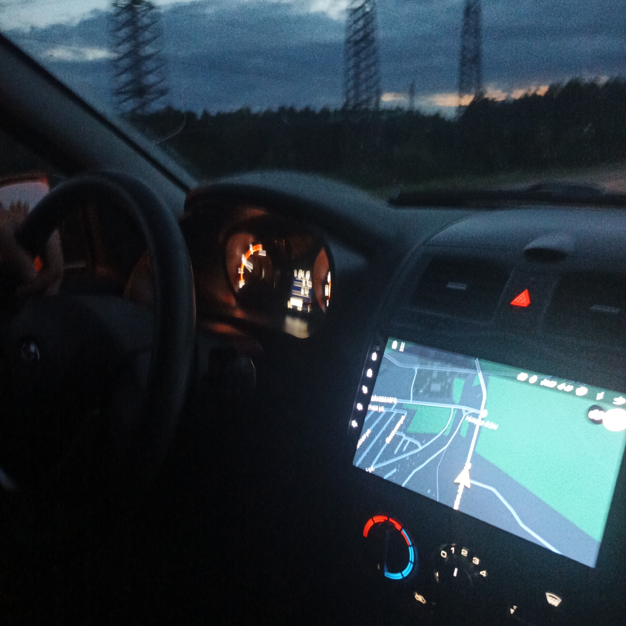 The image is a first-person view from inside a car at dusk. The driver's hand is visible on the steering wheel. The car's dashboard is illuminated, showing the speedometer and a navigation screen displaying a map. Outside the car, through the windshield, tall power lines and a treeline are visible against a darkening sky with hints of orange and yellow from the setting sun. The scene appears to be on the outskirts of a city, likely in Russia given the text on the navigation screen, which includes Cyrillic lettering. The overall impression is of travel during twilight.