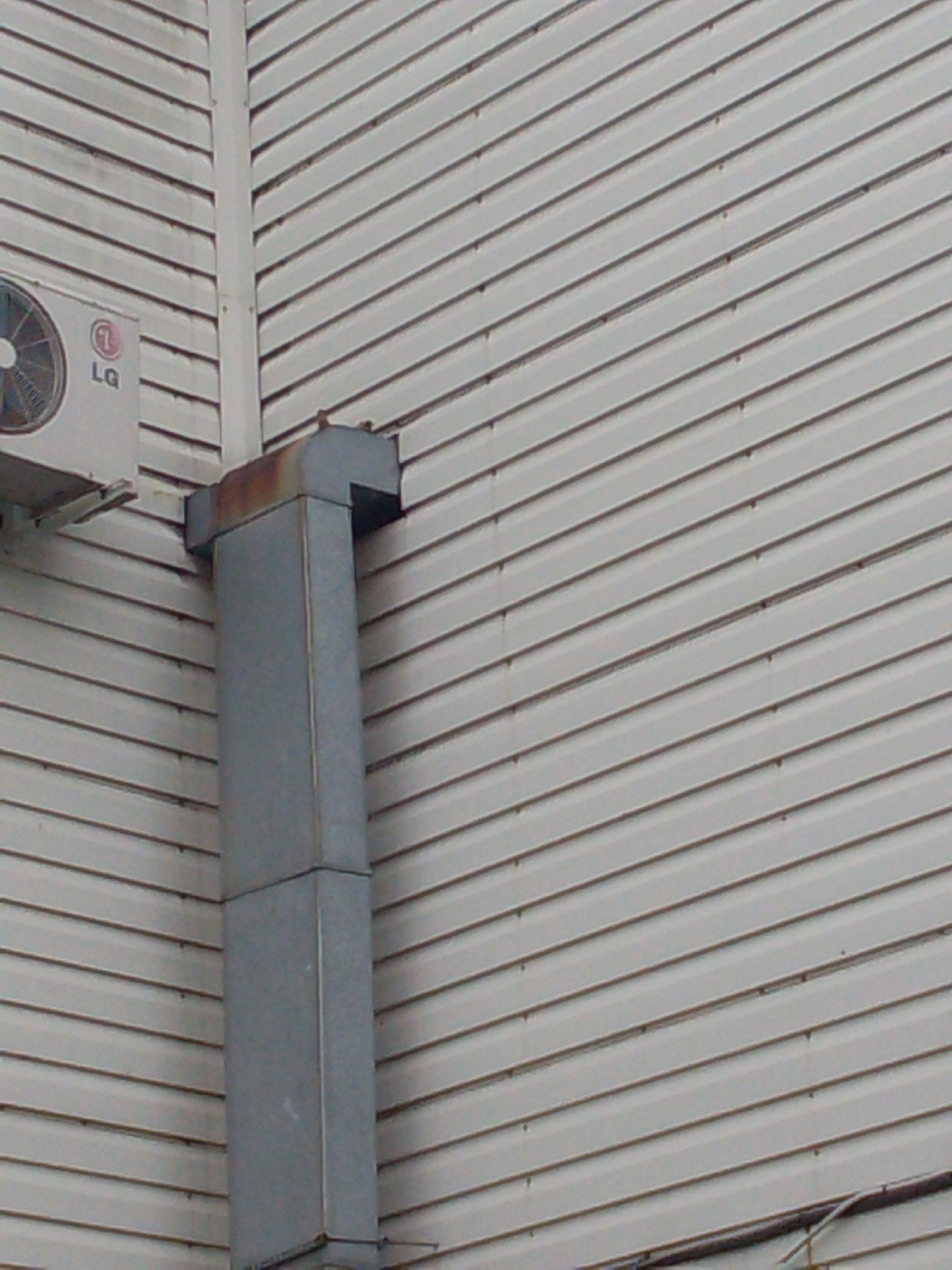 An outdoor shot shows the side of a building with light-colored horizontal siding. A silver-colored air conditioning unit is mounted on the left side of the building, bearing the LG logo. To the right of the air conditioner, a grey metal ventilation pipe is attached vertically to the side of the building. The pipe has a rusty section at the top where it connects to a horizontal piece. The building is situated in a city environment.