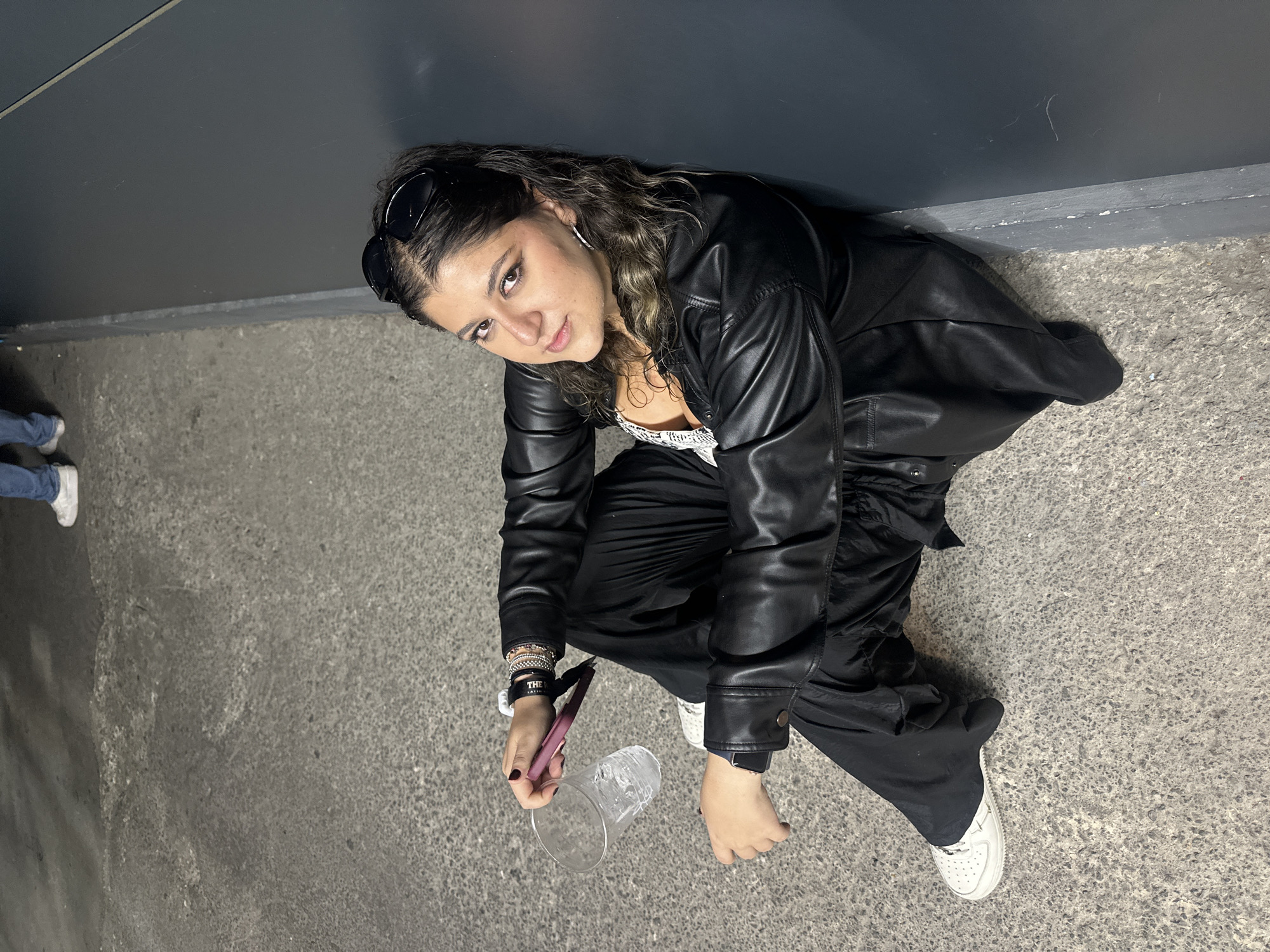 A young woman in a black leather jacket and black pants is crouched on a grey concrete surface next to a dark grey wall. She is looking up at the camera. Sunglasses are perched on her head. She is holding a clear plastic cup and a purple object in her right hand. She is wearing white sneakers. In the lower left corner of the image, the lower legs and feet of another person wearing jeans and white sneakers are partially visible. The location is suggested to be Mexico City, Mexico.