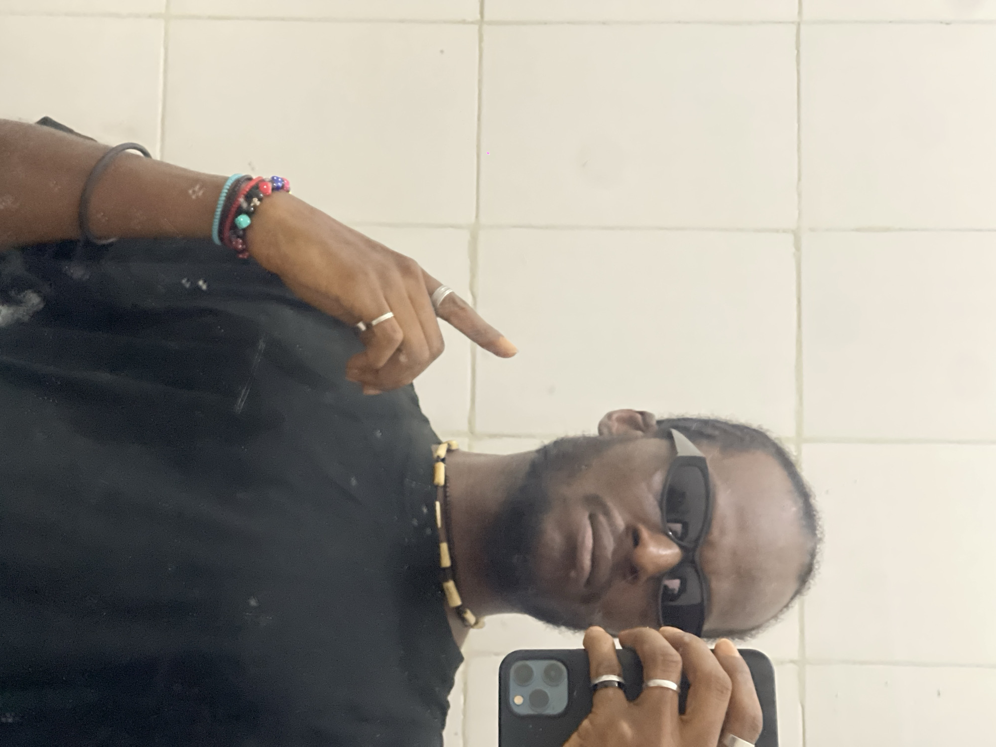 An indoor mirror selfie taken in Jos, Nigeria, captures a dark-skinned male subject. The subject wears dark, rectangular sunglasses, a black t-shirt, and a beaded necklace composed of alternating dark and light cylindrical segments. His short, dark hair is receding, and he has a visible beard.
On the subject's right wrist (appearing on the left side of the frame), multiple beaded bracelets in red, blue, black, and turquoise are visible, along with a dark elastic band. A silver-colored ring is worn on the index finger of this hand, and another on the ring finger; the index finger is raised and pointed upwards. The subject's left hand (appearing on the right side of the frame) holds a dark-colored smartphone, positioned to capture the reflection. The phone's multi-lens camera module is prominently visible. A silver-colored ring is worn on the ring finger of this hand, and a black and silver ring is on the middle finger.
The background consists of a white or off-white tiled wall reflected in the mirror surface, displaying a grid pattern of light-colored grout lines. Minor smudges and specular reflections are evident on the mirror.