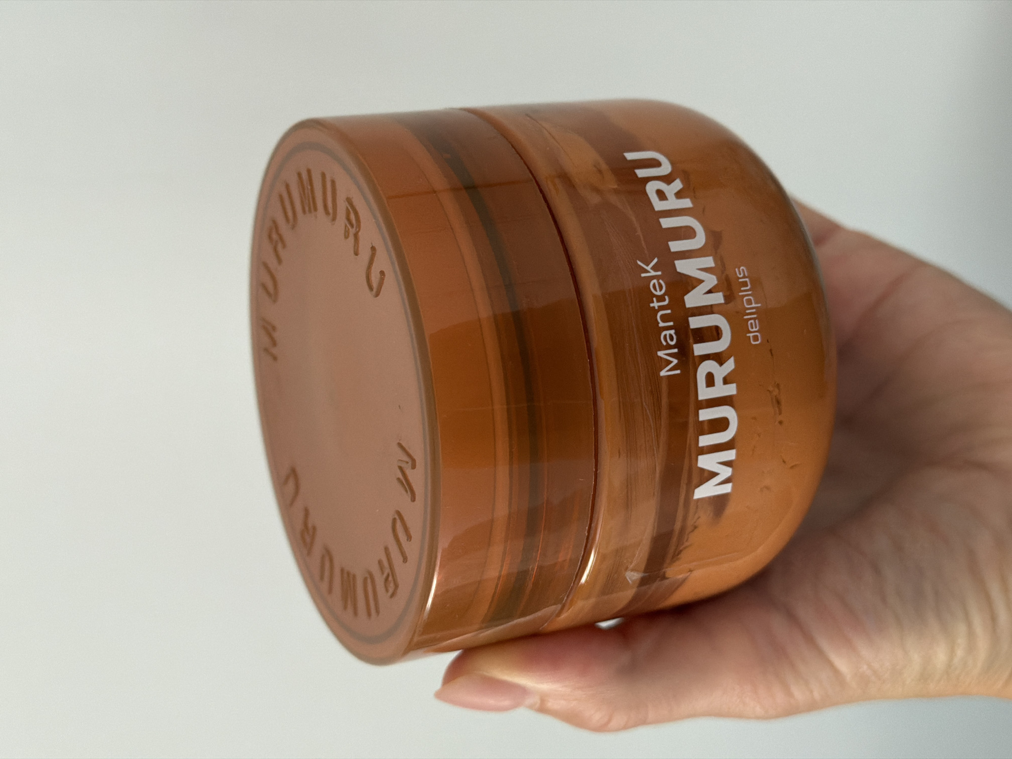 A hand is holding a transparent orange plastic tub of "Mantek MURUMURU" skincare product with the brand name "deliplus" visible below. The lid of the tub has "MURUMURU" embossed in an arc. The background is a plain, light-colored wall, likely indoors. The image is not specific to Finestrat, Spain.
