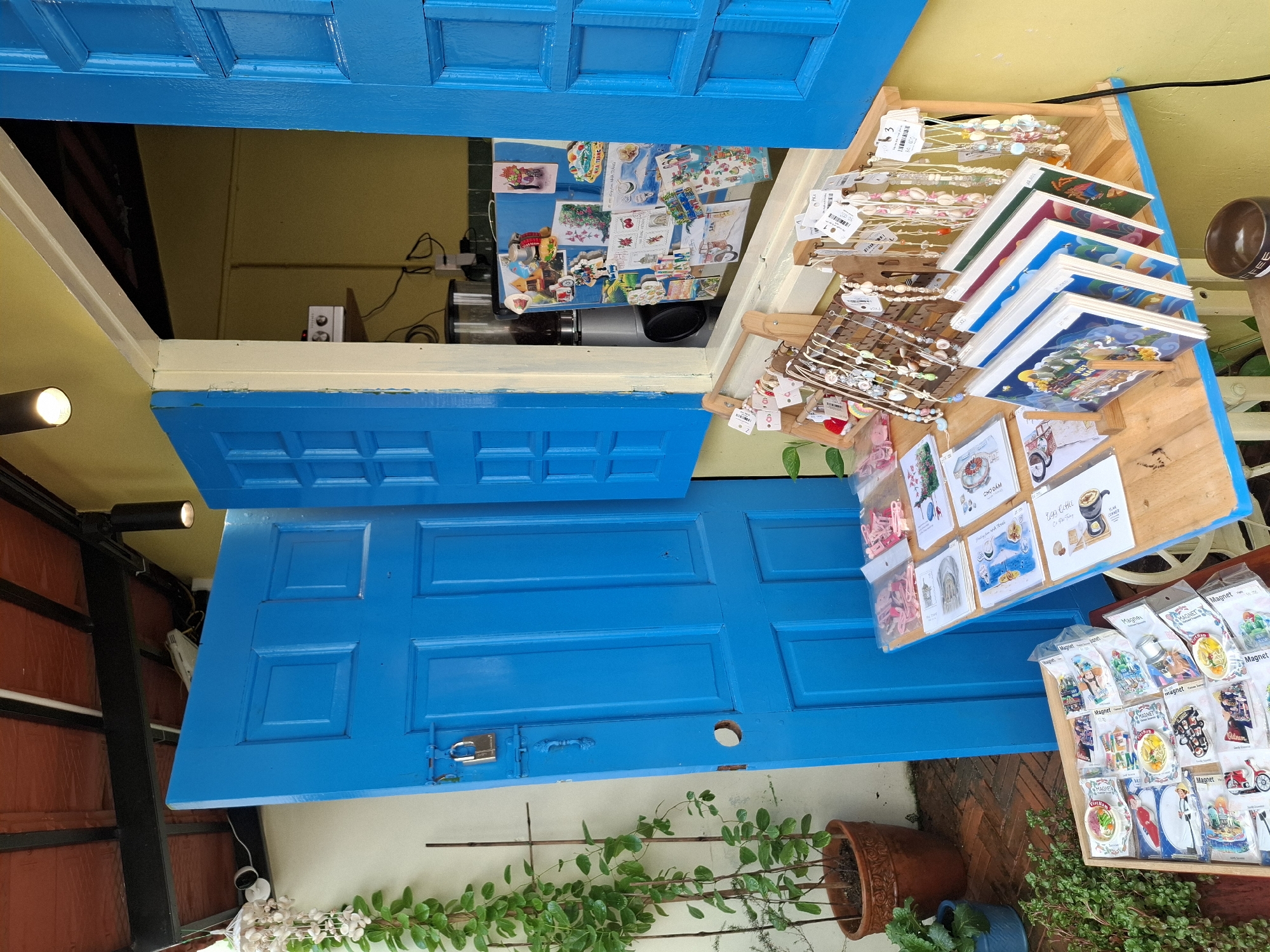 The image depicts a vibrant outdoor scene, likely a shop or stall, in Nha Trang, Vietnam. The focal point is a bright blue, open door, revealing a yellow-walled interior. On the exterior, a table is laden with various small items for sale, including postcards, magnets, and beaded necklaces. The postcards are displayed in a fanned arrangement, showcasing colorful illustrations. A wooden stand holds numerous necklaces, some adorned with seashells. To the left of the door, a potted plant with lush green foliage climbs a trellis. Further left, a blue pot contains another plant. The ground is paved with reddish-brown bricks. In the background, through the open door and a window, a coffee machine and a collection of magnets on a blue surface are visible. The overall impression is one of a charming, colorful, and inviting marketplace stall.