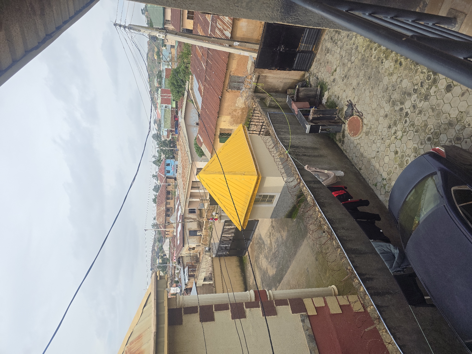 The image shows a view of a residential area in Jos, Nigeria, from an elevated perspective. In the foreground, a dark-colored sedan is parked on a paved courtyard with hexagonal tiles. A concrete wall with barbed wire runs along the courtyard, separating it from other properties. Clothes are hanging on a line attached to the wall. Adjacent to the wall is a small building with a bright yellow, pyramid-shaped roof.
Further back, a black metal gate leads to another property. Beyond these immediate structures, a densely packed neighborhood of various houses with corrugated iron and tin roofs is visible. Some buildings have brightly colored painted facades, while others appear to be constructed with mud or rough plaster. The landscape is hilly, with more houses scattered across the slopes in the background. Utility poles with a network of electrical wires are prominent features in the urban landscape. The sky is overcast with gray clouds.