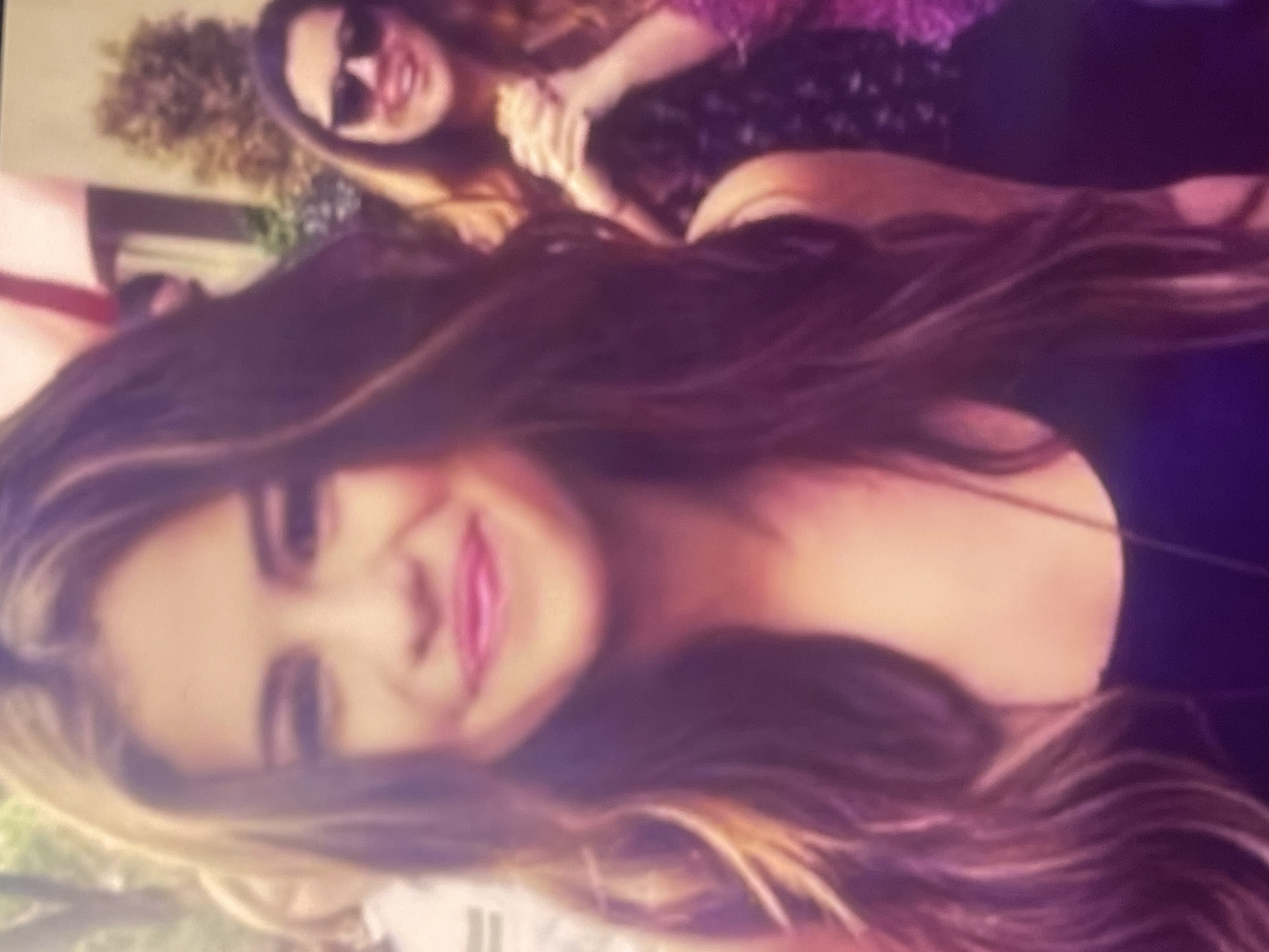 The image, taken in Poltava, Ukraine, captures two women outdoors during the daytime. The photograph is rotated 90 degrees clockwise.
In the foreground, a young woman with long, flowing dark brown hair and fair skin is prominently featured. She is smiling broadly, revealing pink lipstick, and appears to be looking towards the camera. She is wearing a dark-colored top, possibly black or dark blue, with a thin strap visible.
Behind her, slightly out of focus, a second woman is visible. She is wearing dark sunglasses and also smiling. Her hair is long, and she is dressed in a patterned top, which appears to be dark with lighter (possibly purple or pink) floral or abstract details. Her right arm is visible, with her hand gently resting on or holding the arm of the foreground woman, suggesting a friendly or affectionate interaction.
The background shows out-of-focus green foliage, indicating an outdoor setting such as a park or garden. The natural lighting suggests it is daytime. Both women appear happy and relaxed, posing for the photograph. The image has a somewhat soft, slightly blurry quality with warm, desaturated colors, giving it a candid feel. There is no visible text in the image.