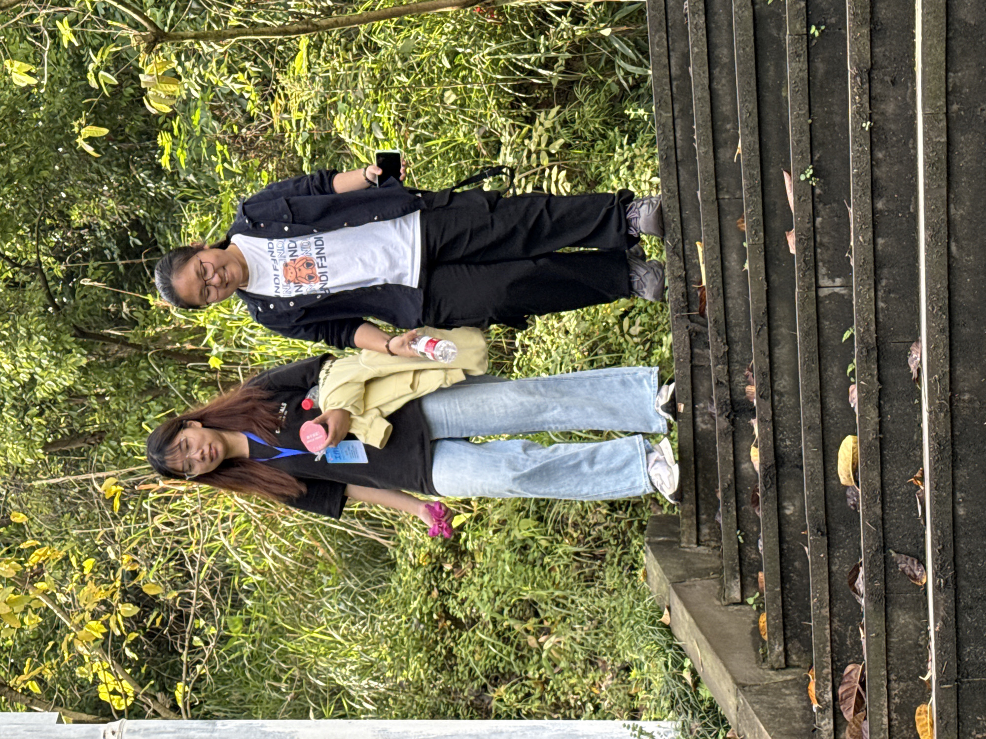 Two young women stand on the side of a concrete staircase in a natural setting. The first woman, closer to the top of the stairs, wears glasses, a white t-shirt with Fendi branding, a black jacket, and black pants. She holds a cell phone in her right hand and a water bottle in her left. The second woman, closer to the bottom of the stairs, wears glasses, a black t-shirt, blue jeans, and white sneakers. She has a yellow sweater draped over her shoulder and holds a flower in her left hand and a water bottle in her right hand. The staircase runs along a hillside covered with vegetation, including trees and various plants. The specific location is Jiangjin, China.