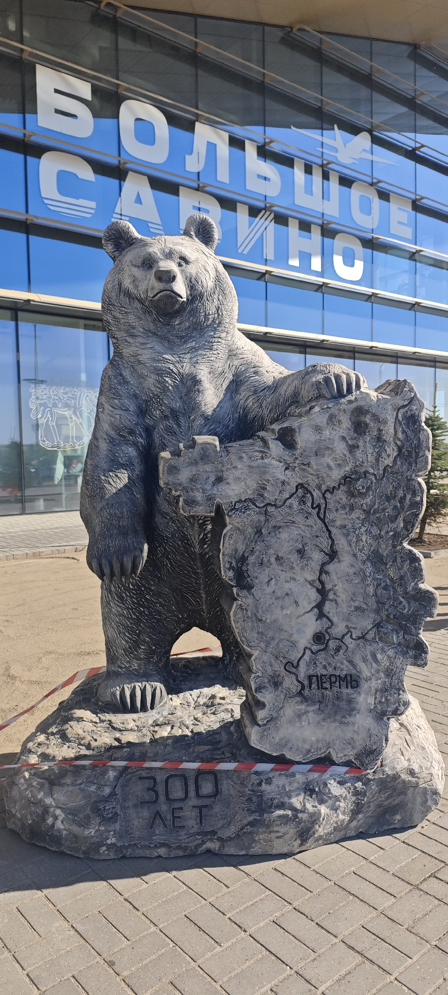 The image shows a bear statue in front of a building in Bolshoye Savino, Russia. The statue is a gray bear standing upright with one paw resting on a stone carving depicting a map with the word "Пермь" (Perm) inscribed on it. The base of the statue has the inscription "300 ЛЕТ" (300 Years). In the background, the building has a glass facade with the inscription "БОЛЬШОЕ САВИНО" (Bolshoye Savino) on it. The scene appears to be outside of an airport terminal in the city of Bolshoye Savino. A red and white barrier tape is around the base.