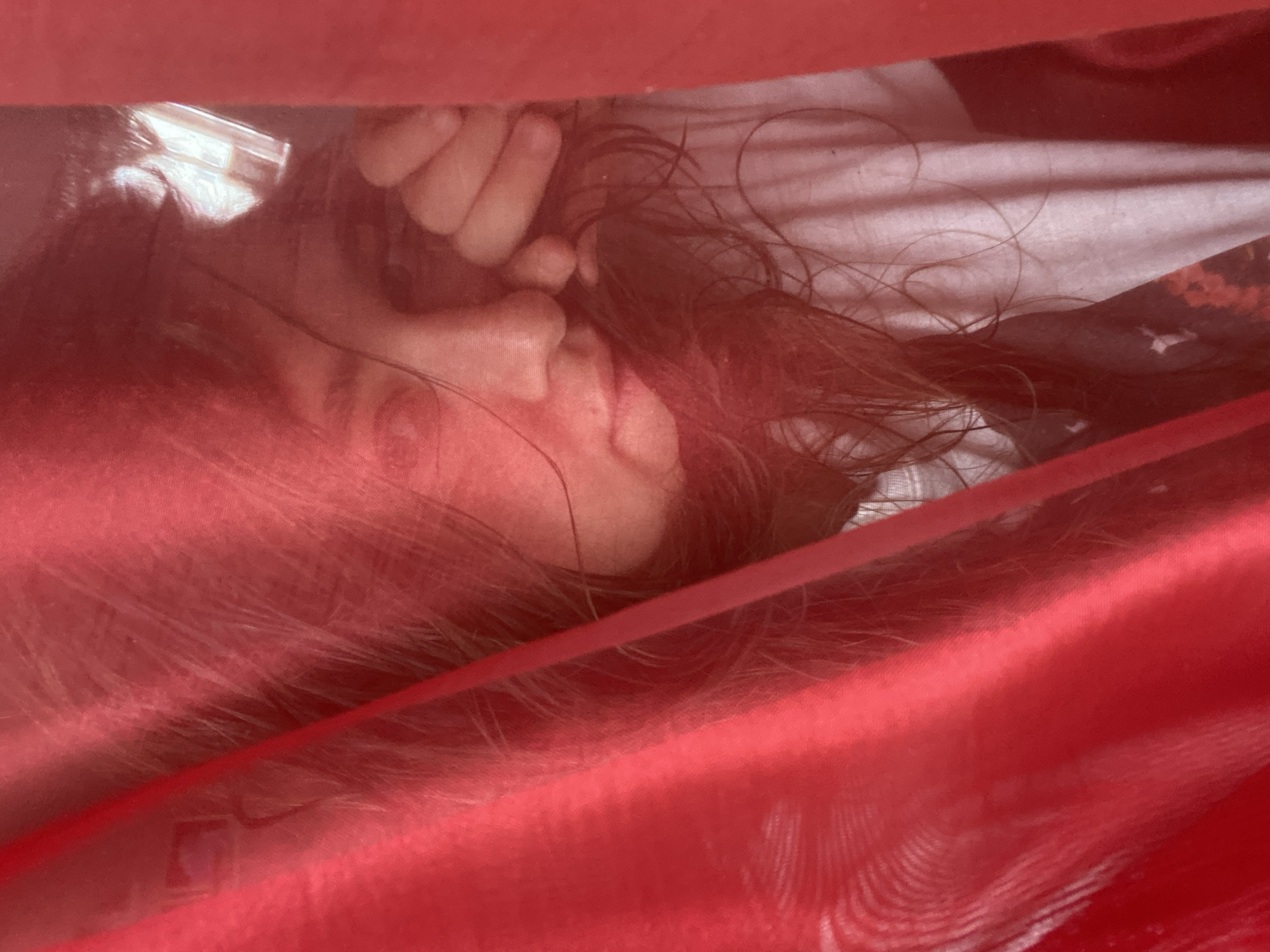A person's face is partially obscured by a translucent red fabric. The person has long, brown hair that falls around their face and shoulders. Their eyes are visible, and one hand is resting near their cheek. They are wearing a light-colored t-shirt. The background appears to be indoors, with a window visible through the fabric, suggesting a residential or indoor setting. There is no visible indication of the city of Perm, Russia.