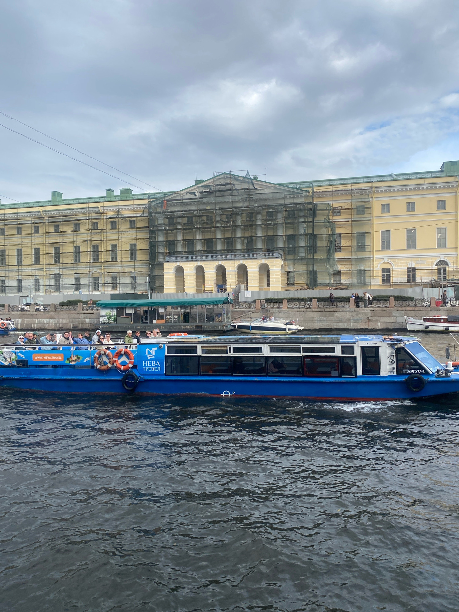 A tour boat, painted bright blue with red trim, cruises down a wide, dark-water canal in Saint Petersburg, Russia. The boat, labeled "НЕВА ТРЕВЕЛ" and featuring the website "www.neva.travel", is filled with passengers, many of whom are looking at the camera or the scenery. The boat's windows are tinted, and a life preserver hangs on its side.
In the background, a grand, yellow building with green trim stands on the embankment. The building is covered in scaffolding, suggesting renovation or construction work. The sky above is overcast with heavy gray clouds, characteristic of Saint Petersburg's often moody weather. Several other boats are visible on the canal, including a smaller white motorboat to the left and a white boat with a red stripe to the right, further down the canal. The overall scene depicts a typical tourist experience in the historic city.