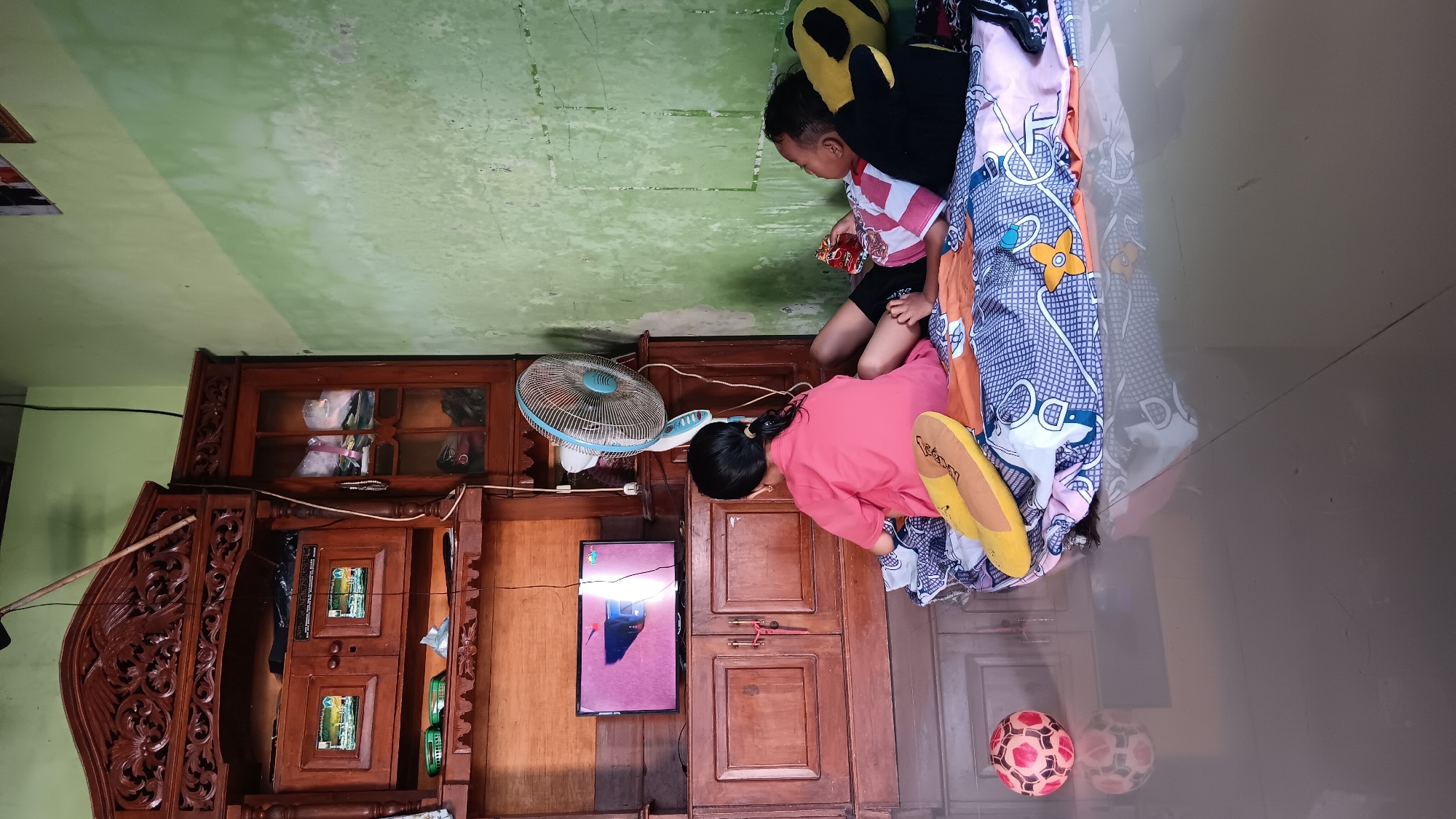 In Sonobekel, Indonesia, two children are observed lying on the floor, engrossed in watching television. A young girl in a pink shirt is seated facing the screen, while a younger boy lies beside her, propped up by stuffed pillows. The scene takes place in a room with a wooden cabinet dominating one side, housing a television set and various items. A blue oscillating fan is positioned near the cabinet. The floor is tiled and reflects the room's contents. A colorful soccer ball rests on the floor to the left of the children. The walls are painted in a pale green hue.