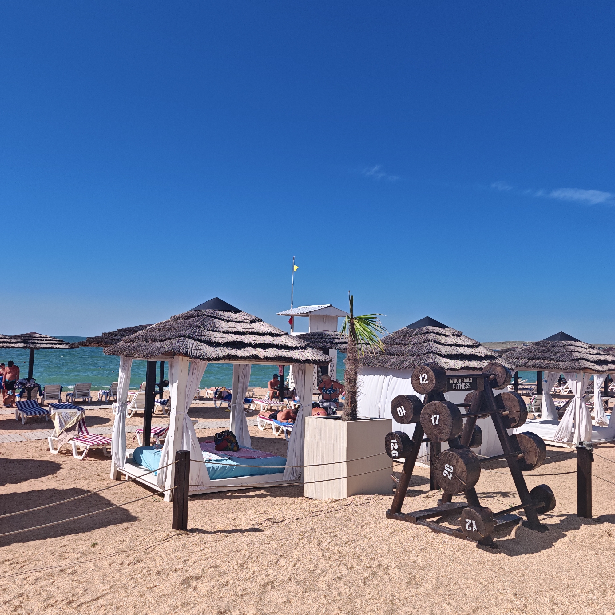This is a full-shot, eye-level, outdoor photograph of a beach scene. The sky is a clear, bright blue, taking up the top portion of the image. The foreground and midground are occupied by a sandy beach with several structures and objects.
In the midground, there are several thatched-roof cabanas. Some are open, revealing lounge beds with white curtains. People are visible relaxing in and around these cabanas. In front of some of the cabanas are lounge chairs and umbrellas.
To the right of the image, a wooden rack holds several large, circular weights, resembling dumbbells, with numbers painted on them (e.g., 10, 12, 12.6, 17, 20, 21). The text "WOODSMAN FITNESS" is also visible on one of the weights. A small palm tree is situated next to the weight rack, in front of a white tent-like structure.
Further in the midground, the turquoise sea is visible with gentle waves. A lifeguard tower with a yellow flag is situated on the beach between two cabanas. Other beachgoers can be seen in the distance, some in the water and some on the sand. The overall impression is a resort-like beach environment on a sunny day.
The location is identified as Мысовское сельское поселение, Ukraine.