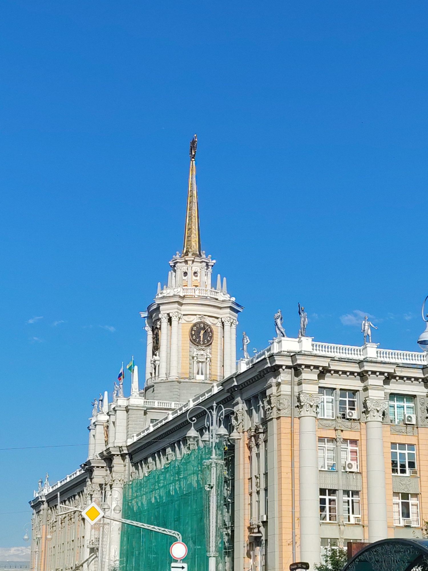 The image captures a view of the Yekaterinburg City Hall, a prominent Stalinist-era building located at 108 Lenina Avenue, Yekaterinburg, Russia. The architectural style is characterized by its imposing facade, adorned with classical columns, statues, and a striking central tower topped with a golden spire. The building is partially covered by green scaffolding and netting, suggesting ongoing renovation work.
In the foreground, a street sign with a yellow diamond symbol and a circular traffic sign are visible. The sky above is a clear, vibrant blue, with a few wisps of clouds. The overall scene conveys a sense of the city's grandeur and historical significance, juxtaposed with contemporary urban development and maintenance.