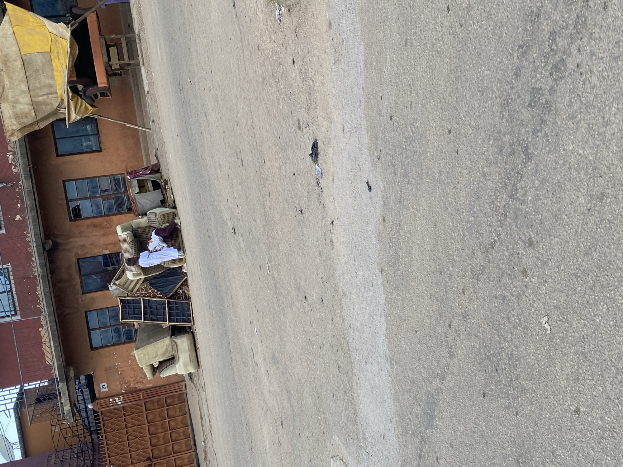 In Jos, Nigeria, in front of an orange building, a pile of discarded furniture, including sofas and chairs, sits on the side of a paved road. Two individuals are visible near the furniture pile. One person, dressed in a white garment, is standing and appears to be interacting with the other person who is seated and partially obscured by the furniture. A large brown metal gate is to the left of the scene, and a makeshift awning covered with tarpaulin is to the right. The road surface shows signs of wear and is littered with debris.