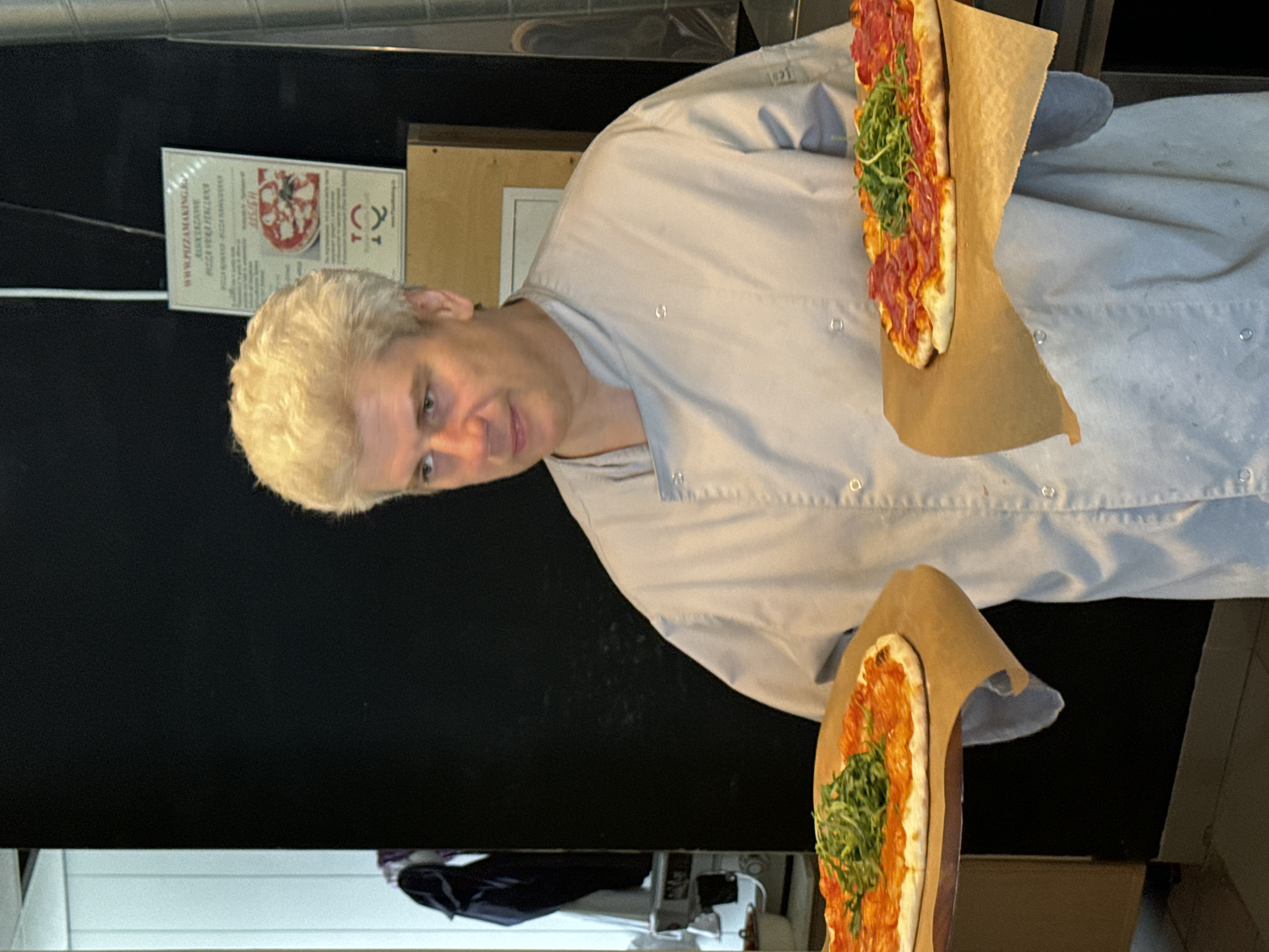 A man wearing a light gray chef's coat holds two pizzas. The pizza on his left has a red sauce and a topping of green arugula. The pizza on his right has a red sauce, pepperoni, and a topping of green arugula. The man is looking slightly to his left. Behind him, on a black wall, is a sign with text and an image of a pizza. The sign is in a frame. The man and pizzas are in the foreground. To the left of the man, a doorway or opening reveals a glimpse of a white wall and a black garment hanging. To the right of the man, a silver metallic structure is visible. The man appears to be in a restaurant or kitchen setting. The location is in the city of Saint Petersburg, Russia.