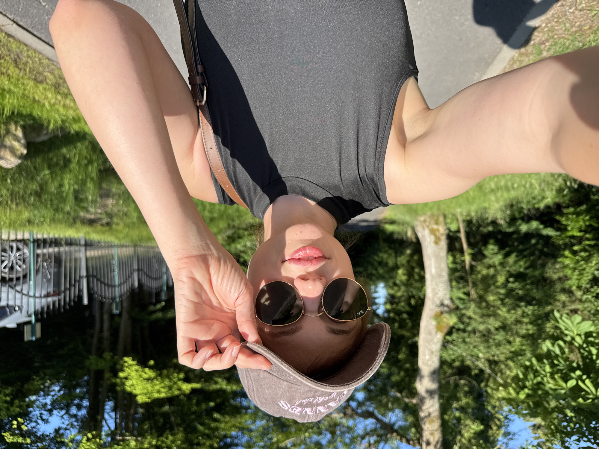 A young woman with fair skin, wearing a dark sleeveless top, a brown baseball cap, and round sunglasses, is taking a selfie outdoors. She is holding the brim of her cap with her right hand. The background shows a paved path, lush green grass, trees, and a black metal fence with a parked car visible behind it. The lighting suggests it is daytime, with sunlight casting shadows. The location is identified as Szklarska Poręba, Poland.