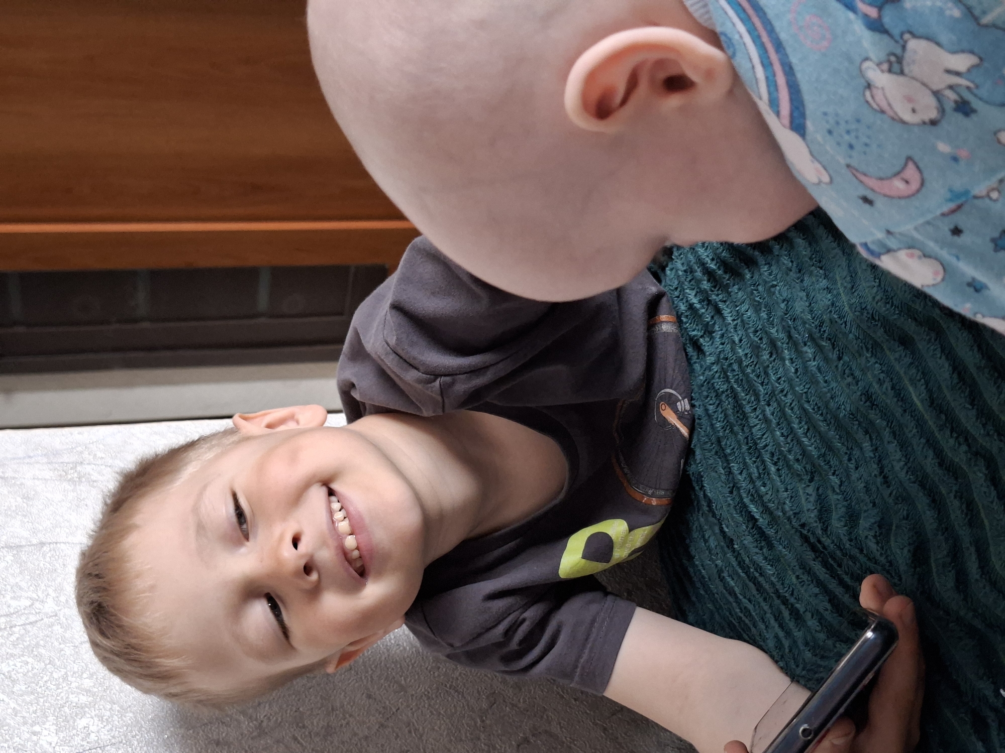 A close-up shot depicts two children, one older and one younger, engaged in an interaction. The older child, a fair-skinned boy with light blond hair, is looking at a mobile phone held in his hands. He is smiling and showing his teeth. He is wearing a gray t-shirt with graphics on it. The younger child, presumably a baby, is partially visible with a pale complexion and very little hair. The baby is wearing a light blue patterned garment. The two are positioned over a textured, dark green surface that looks like a blanket. The backdrop includes a dark wooden surface above a light gray and white floor. The location is Unknown, Unknown.