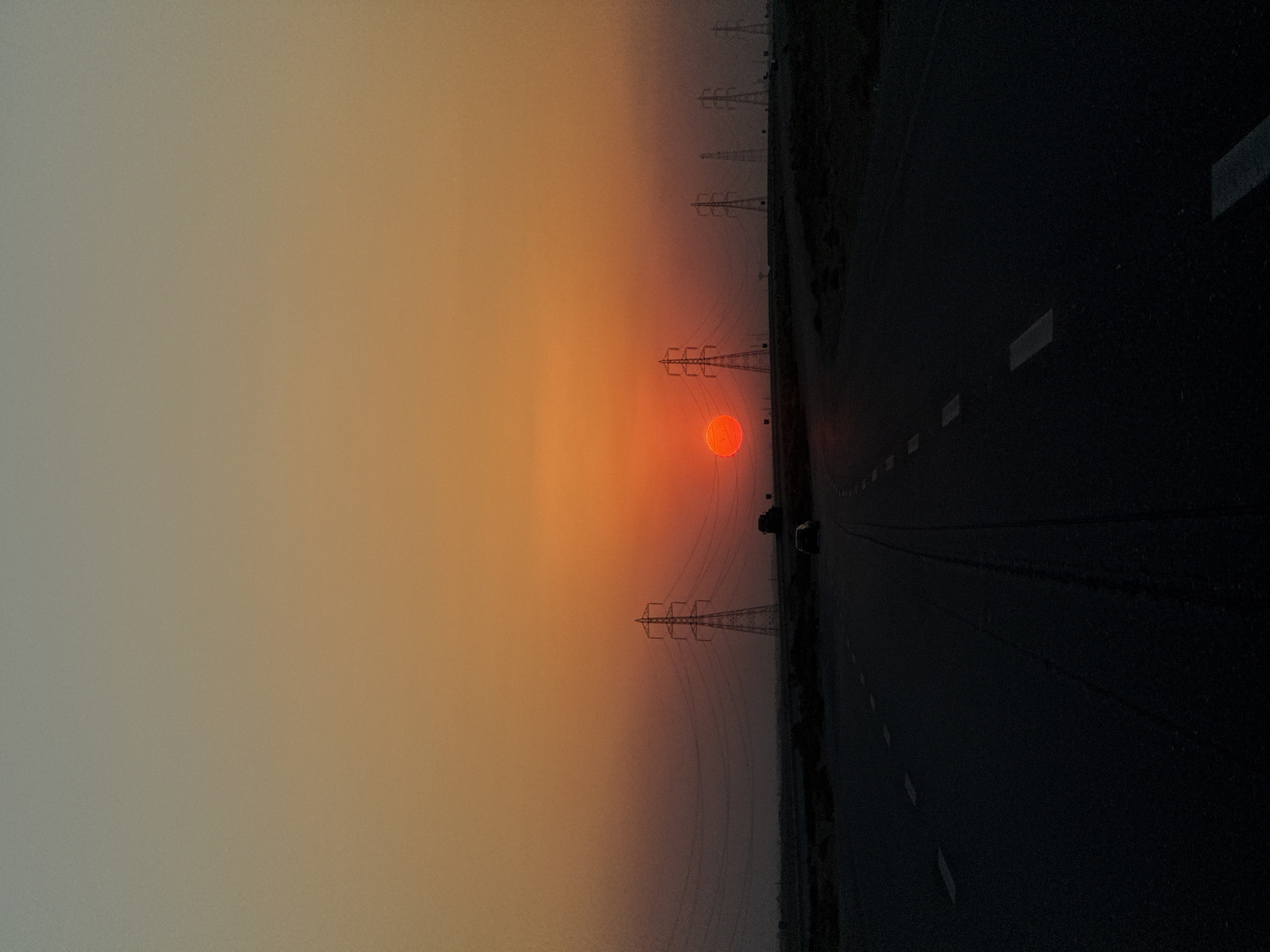 The image shows a highway at sunset. The sun is a vibrant orange disk near the horizon, casting a warm glow across the sky. Power lines stretch across the landscape, silhouetted against the colorful sky. The highway is dark and appears wet, with white lines marking the lanes. A small vehicle is visible on the side of the road. The overall mood is serene and contemplative, with the setting sun and empty highway evoking a sense of journey and transition. The time of day is clearly sunset. The weather appears to be clear, with a hazy sky contributing to the softened colors of the sunset. No text is visible in the image.