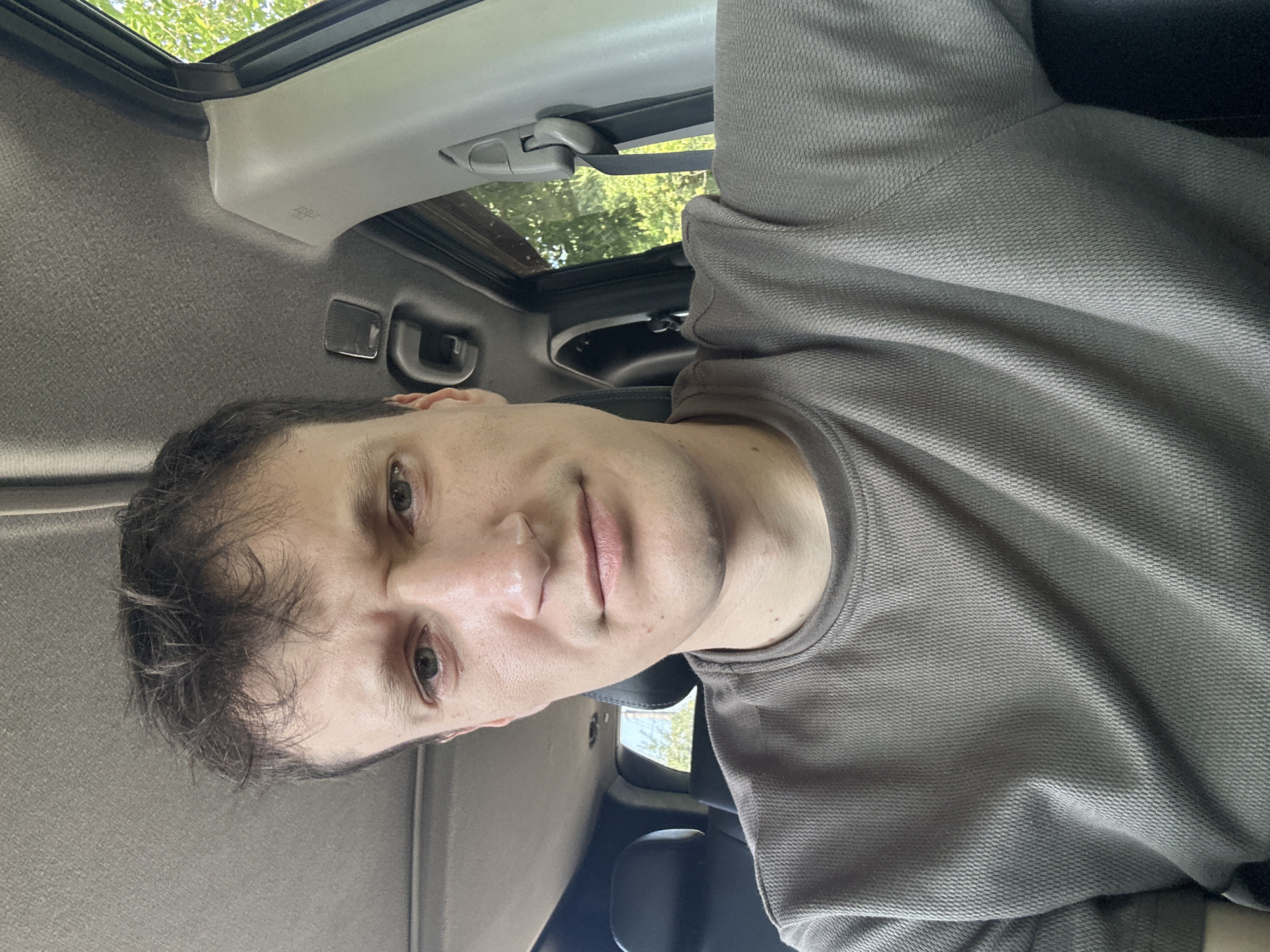 A close-up, selfie-style shot shows a young adult male with light brown hair and a gray t-shirt, seated inside a vehicle. The man's face is the primary focus, with his brow furrowed slightly and his eyes looking towards the camera. The interior of the car is visible, including the ceiling, a door frame, and the headrests of seats. Outside the car window, trees and foliage can be seen, suggesting a daytime setting in a green, possibly rural or suburban, environment. There is no specific indication of the location being in Makiv, Ukraine, or any city.