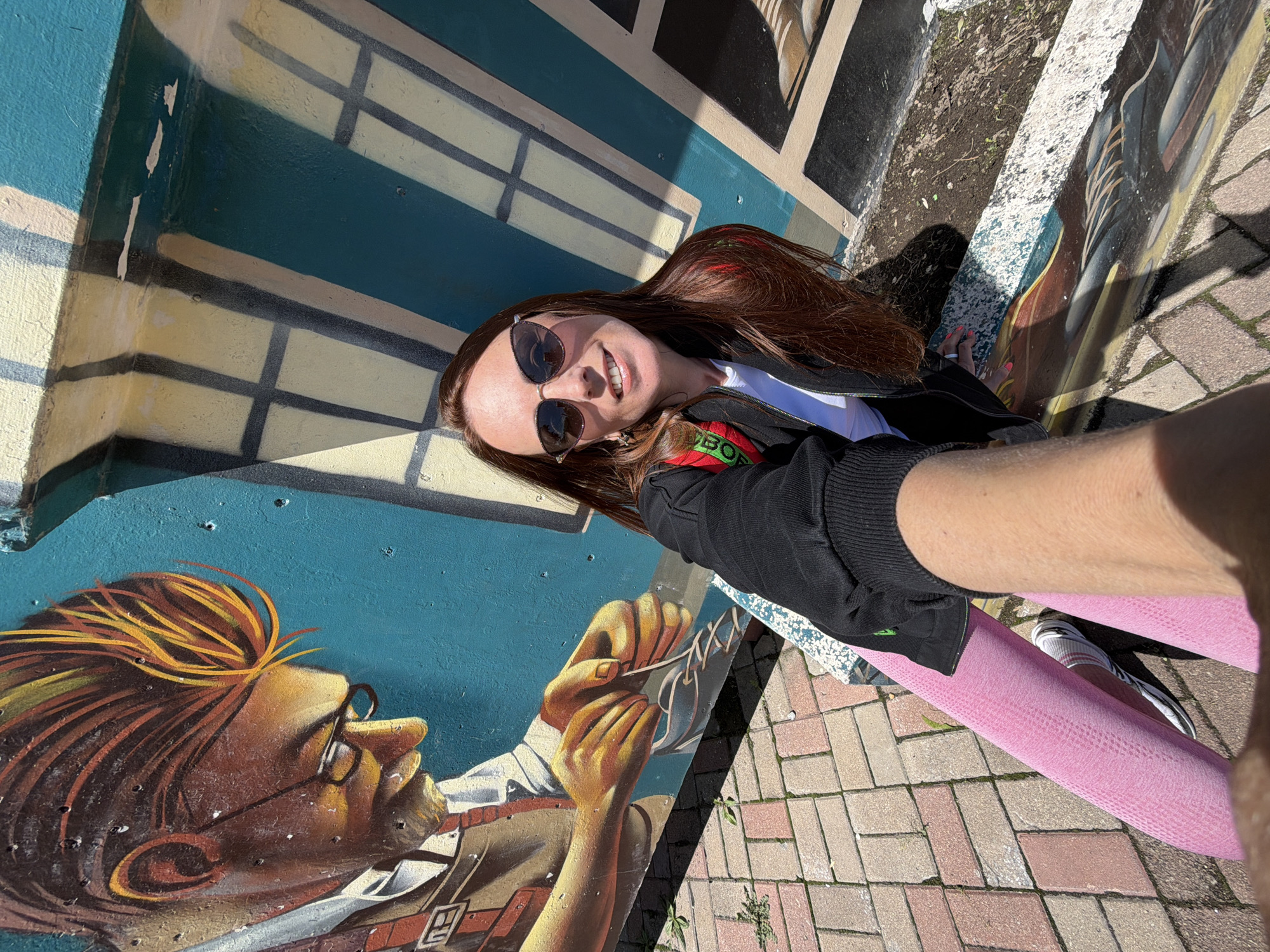 The image shows a woman taking a selfie in front of a colorful mural. The mural depicts a close-up of a person's head and hands, with a teal background and a stylized window pattern. The woman is wearing sunglasses, a black jacket over a white shirt, pink leggings, and white sneakers. Her right arm is extended to capture the selfie. The ground is paved with red and tan bricks. The scene appears to be outdoors on a sunny day, with shadows cast by the sun. There is no visual information in the image that suggests the location is Birobidzhan, Russia.