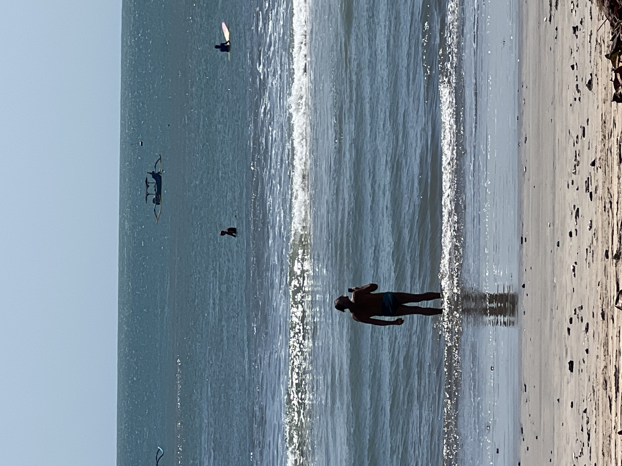 A man stands in the shallow water of Kuta Beach, Indonesia, facing away from the camera, holding a phone to take a photo. The ocean is calm with gentle waves. Further out, a traditional Indonesian fishing boat with outriggers is visible, along with two surfers on their boards. The sky is clear and blue. The foreground shows a sandy beach littered with small stones and debris.