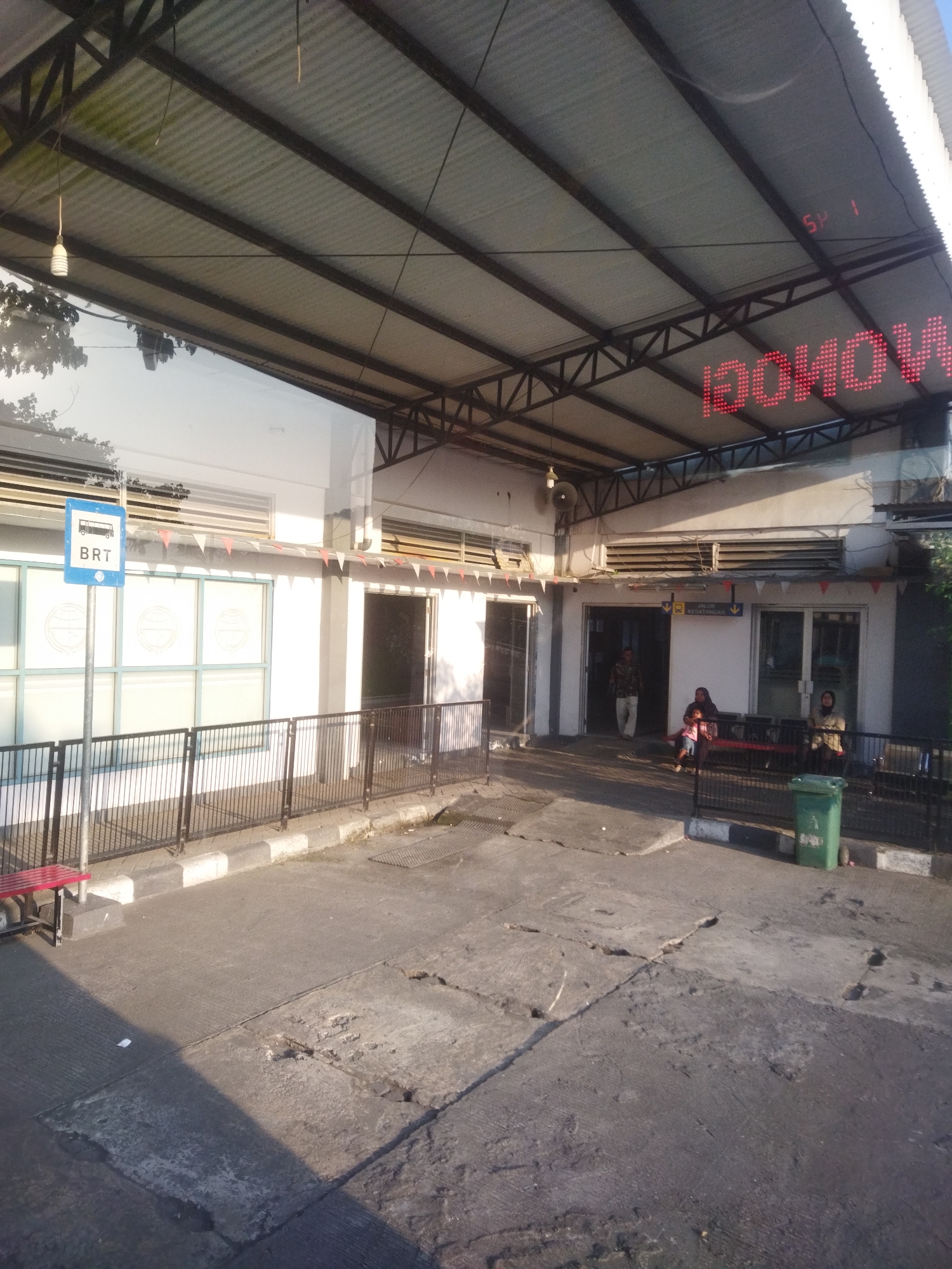 The image depicts a bus stop area, likely within the city of Kaliancar, Indonesia, given the context. The scene is partially covered by a corrugated metal roof supported by a metal frame. To the left, a blue sign with "BRT" and a bus icon indicates the bus rapid transit service. Several glass-paneled windows are visible on the white building behind the bus stop sign. A black metal fence runs along the front of the building.

In the background, the building has entrances, and some individuals are present. A man in a patterned shirt and light pants is walking towards one of the entrances. Further to the right, a woman with a child is sitting on a red bench, and another woman is seated on a bench nearby. A green trash can is located near the right edge of the image.

The ground in the foreground is paved with concrete, showing signs of wear and cracking. Sunlight casts shadows across the area, suggesting it is daytime. Above, a red LED sign displays text that appears to be partially obscured or distorted, but a portion of it reads "BOHOWA" and "SP 1." Small triangular flags are strung along the edge of the roof above the building's facade. Red benches are positioned along the bus stop area.
