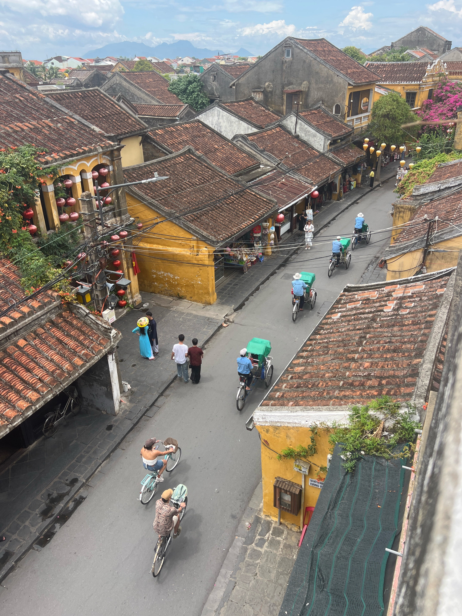 This is an aerial, high-angle shot of a street scene in Hoi An, Vietnam, with a focus on the traditional architecture and daily life. The image captures a narrow street lined with tightly packed, yellow and muted-colored houses with distinctive terracotta tiled roofs. Many of the houses have open fronts, revealing shops or living spaces. Red lanterns are visible hanging from some of the buildings, suggesting a festive atmosphere or a typical street decoration in this historic town.
Several people are depicted on the street, engaging in various activities. In the foreground, two people are riding bicycles, one with a basket on the front. Further up the street, three cyclos (three-wheeled bicycle taxis) are moving, each carrying a driver and a passenger or cargo covered by a green tarp. One cyclo driver is wearing a white hat. In the middle ground, two people are walking away from the camera, and a person in a blue dress and a conical hat is standing still. A group of three people are standing and conversing on the sidewalk. In the distance, more houses and buildings extend into the background, with a hazy mountain range visible on the horizon under a partly cloudy sky. The overall impression is of a vibrant and bustling urban environment with a strong sense of history.