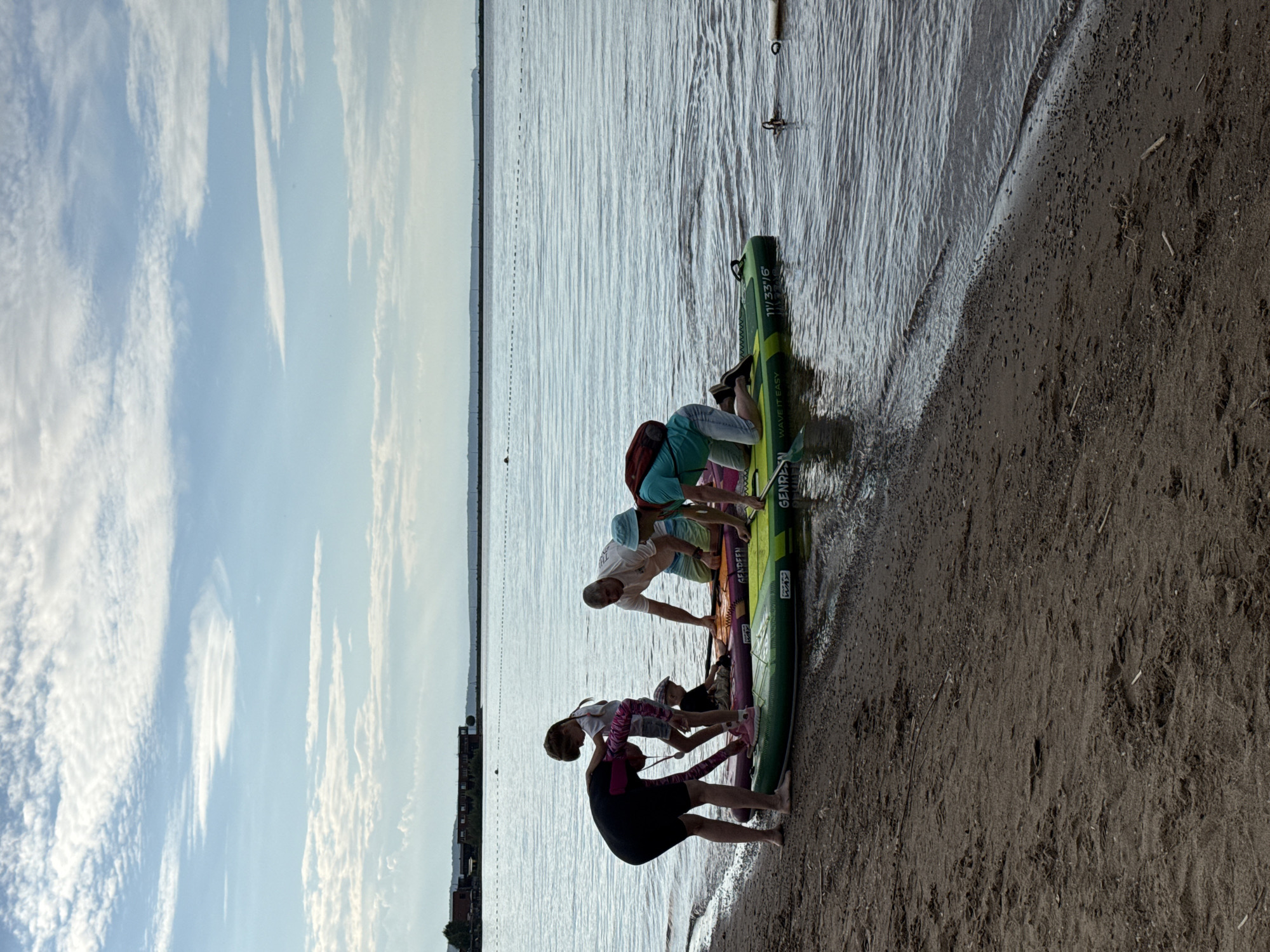 The image depicts a group of people preparing for or disembarking from paddleboarding on a body of water, likely the Kama River, near the city of Chaikovsky, Russia. The scene is set on a sandy beach with the water lapping at the shore. A variety of paddleboards are present, some partially submerged in the water and others on the sand.
In the foreground, on the left, a person in a dark full-body swimsuit with pink accents is bent over, interacting with a young girl who is standing and holding onto the swimsuit. Another child, wearing a cap, is sitting on a paddleboard in the middle. To the right of these figures, a man in a white t-shirt and shorts is kneeling on a paddleboard, and further to the right, another person in a teal shirt and shorts is also kneeling on a paddleboard. The people appear to be engaged in a group activity related to paddleboarding.
The environment is characterized by a calm, rippled body of water, a sandy beach, and a cloudy sky with patches of blue. In the distance, across the water, a shoreline is visible with some buildings and structures, possibly part of the industrial or residential area of Chaikovsky. A breakwater or barrier line is also discernible in the water, extending towards the horizon. The lighting suggests it is daytime, likely late afternoon or early evening due to the soft quality of the light.