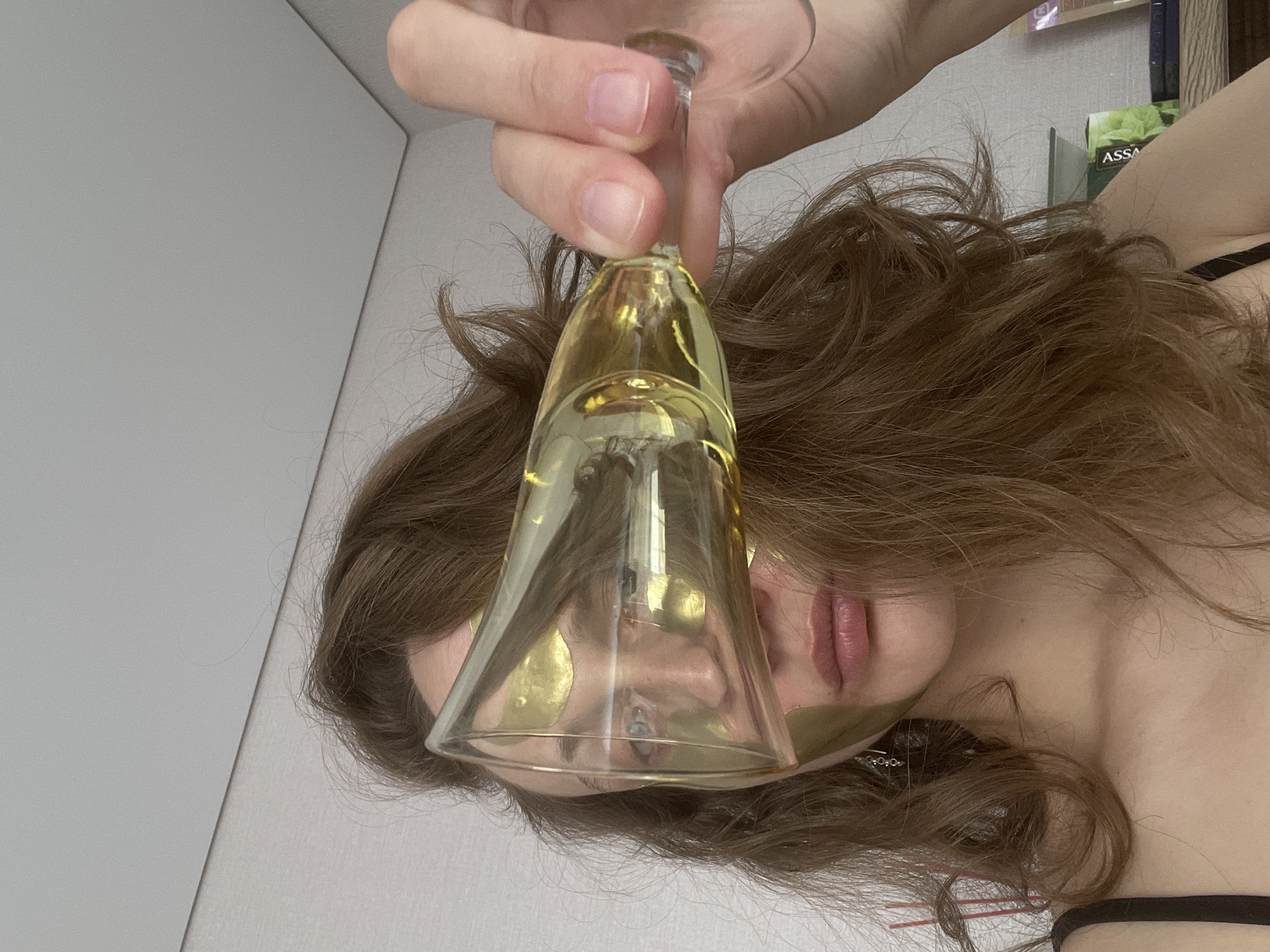 An indoor, close-up image, rotated ninety degrees clockwise, captures a person engaged in a moment of relaxation and self-care. The central subject is a person, likely female, whose face is partially obscured by a clear stemmed glass, possibly a champagne flute, held by her hand. The glass contains a yellowish liquid or has a golden tint, and is positioned directly in front of her face.
The person has wavy, reddish-brown hair that flows around her head. She is wearing gold-colored under-eye patches, indicating a beauty or relaxation treatment. Her full, light pink lips are visible, along with a glimpse of her eye through the glass. A black bra strap is visible on her shoulder.
The background is minimalistic, featuring light gray walls or a ceiling. In the upper right corner, shelves hold various objects, including what appears to be a box or package with the text "ASSA" visible on its side. The scene suggests a private, calm setting, possibly at home in Podshivalovo, Russia, where the individual is unwinding with a drink and a beauty treatment.
