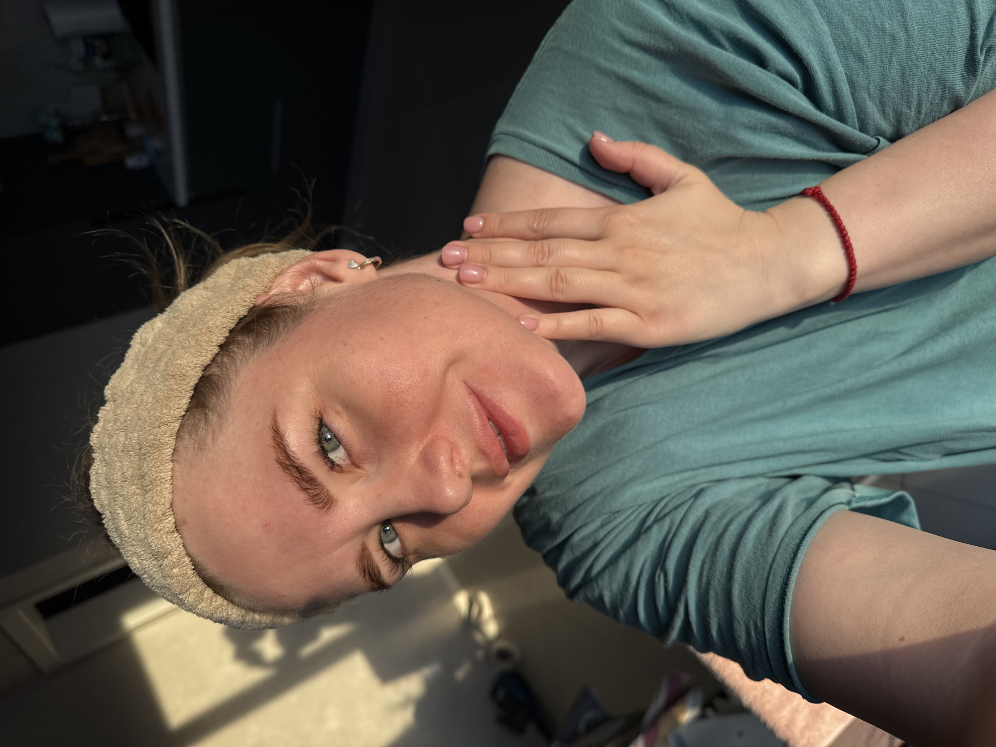 A light-skinned person is facing the camera at an angle. They have light blue eyes, a tan headband, and a small heart-shaped earring. They are wearing a teal-colored t-shirt and have a red string bracelet on their left wrist. Their left hand is raised near their neck. Sunlight illuminates part of the scene. The location is unknown.