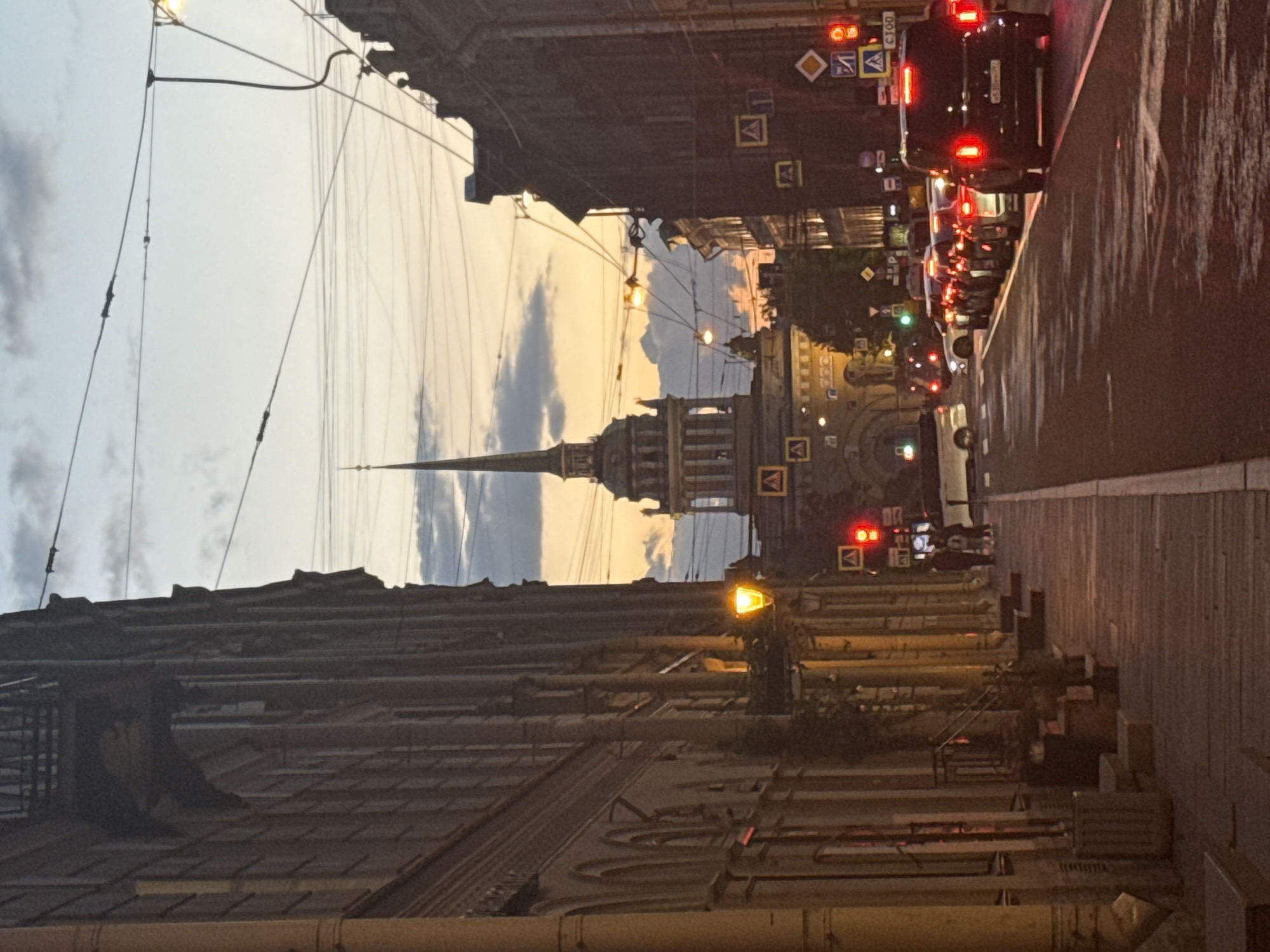 The image captures a street scene in Saint Petersburg, Russia, at twilight or dawn. The perspective looks down a wide, wet, and reflective street towards the iconic Admiralty Building, whose tall, golden spire dominates the middle distance against a soft, gradient sky.
On both sides, the street is flanked by multi-story, classical European-style buildings, their facades partially illuminated by artificial light. Overhead, numerous power lines, likely for trams or trolleybuses, crisscross the sky.
The street itself is busy with traffic. A queue of cars is visible, mostly on the right side of the road, with many red taillights glowing brightly, suggesting they are either stopped at a traffic light or moving slowly. Several traffic lights are visible, some red and some green, regulating the flow. Streetlights along the road cast warm, yellow light onto the scene, creating reflections on the damp asphalt.
Notable details include:
* **Time of day:** The warm hues in the sky, combined with active streetlights and vehicle lights, indicate dusk or dawn.
* **Weather:** The wet appearance of the road suggests recent rain or damp conditions.
* **Location cues:** The distinctive spire identifies the landmark as the Admiralty Building, a central feature of Saint Petersburg.
* **Visible text:** On various traffic signs, symbols for speed limits, crossings, and other regulations are present. One car's rear shows characters "CTOO," possibly on a sticker or partial license plate. A speed limit sign shows "40."
The scene conveys a bustling yet atmospheric urban environment, characteristic of a major European city in transitional lighting.