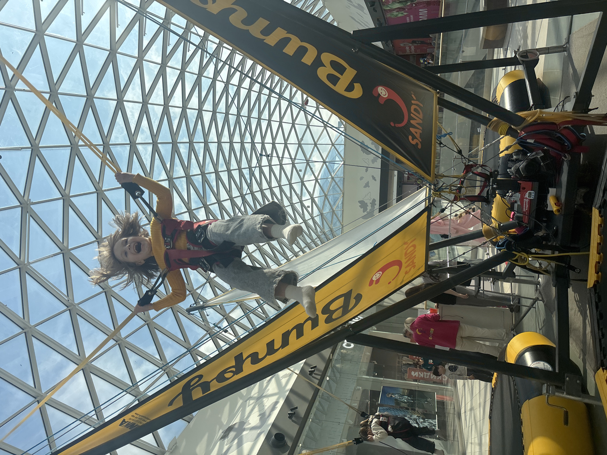 The image displays an active indoor scene at a "Bumphy Sandy" bungee trampoline attraction, located within a large shopping mall in Perm, Russia. The most prominent feature is a child, approximately 6-10 years old, suspended mid-air in the center of the frame, caught in the act of bouncing. The child, with blonde hair flung upwards, wears a yellow long-sleeved top, grey trousers, and white socks, secured by a red and black harness attached to yellow bungee cords extending towards the high, glass-domed ceiling. The child's mouth is open, indicative of shouting or laughter.
The "Bumphy Sandy" attraction consists of a multi-station setup. Two large, upward-sloping black banners, prominently displaying "Bumphy" in yellow Cyrillic script and a red "Sandy" logo featuring a smiling, stylized character, frame the central activity. At the base of the attraction, various mechanical components, ropes, and yellow and black inflatable elements are visible. Equipment bears the labels "Quattro elementi" and "Русский аттракцион" (Russian attraction).
To the left, another child, dressed in a white long-sleeved top and dark pants, is partially visible on an adjacent trampoline station, also wearing a harness. On the ground level, behind a counter associated with the attraction, an adult female wearing a pink hoodie emblazoned with the "Sandy" logo is present, appearing to supervise or operate the ride. Beside her, a young boy in a striped shirt observes the scene. Additional adults are partially visible in the background.
The environment is a spacious, brightly lit interior, characterized by an expansive, geometric glass roof structure allowing natural light to illuminate the area. The background features glass storefronts of retail shops, some with visible Cyrillic text, confirming a commercial mall setting. The floor is clean and reflective.