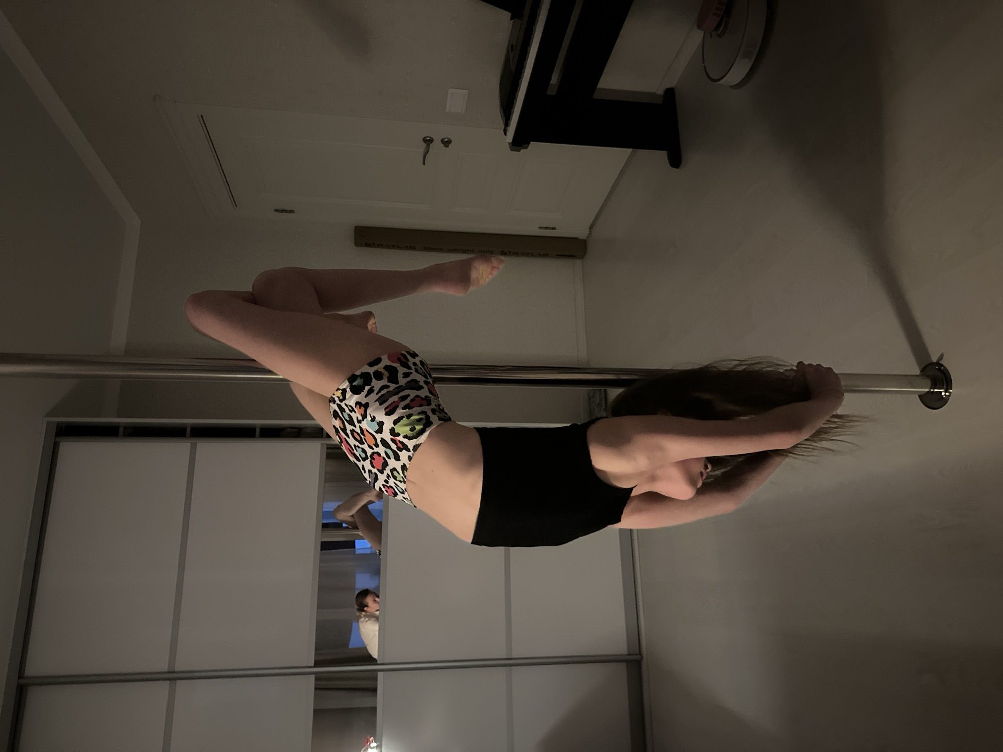A woman wearing animal-print shorts and a black top is hanging upside down from a horizontal metal pole. Her legs are bent, and she is holding the pole with her hands. The setting appears to be indoors, possibly in a residential space with a sliding-door closet in the background. A reflection of a person is visible in the closet mirror. The room also contains a piano and a box. The precise location is unknown.