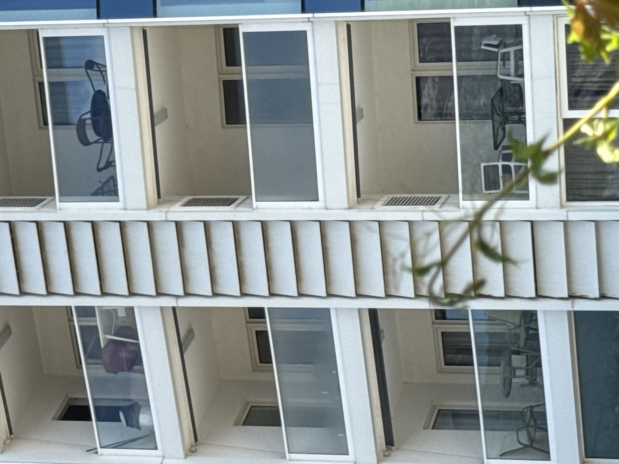 The image shows the balconies of a multi-story building. Each balcony has a glass door and appears to be furnished with chairs and tables, though the details are somewhat obscured by reflections and the angle of the shot. Some balconies have outdoor furniture, suggesting they are part of apartments or hotel rooms. In some of the reflected images within the glass doors, chairs and tables are visible, indicating common outdoor seating arrangements. The building itself has a modern architectural style with prominent vertical elements. The time of day appears to be daytime, with natural light illuminating the scene. The sky is not directly visible, but the lighting suggests it is not overcast. The overall setting evokes a residential or hospitality environment. There is no visible text in the image.
