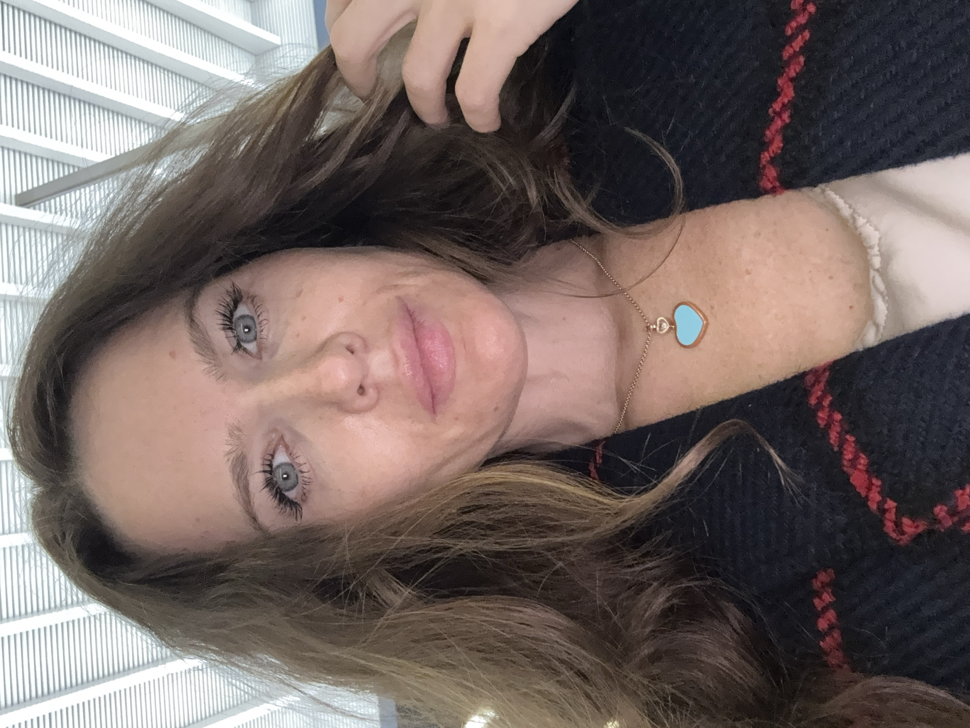A close-up, head-and-shoulders selfie of a woman with light brown, wavy hair is presented. She has light blue eyes and freckles on her face. She is wearing a dark blue and red patterned jacket over a light-colored top. A rose gold chain necklace with a light blue heart-shaped pendant is visible around her neck. Her right hand is near her hair, slightly obscuring the right side of her face. The background appears to be an indoor setting with a slatted ceiling structure, possibly a modern building with a distinctive architectural element. The lighting suggests an artificial light source. There is no indication of the image being located in Moscow, Russia.