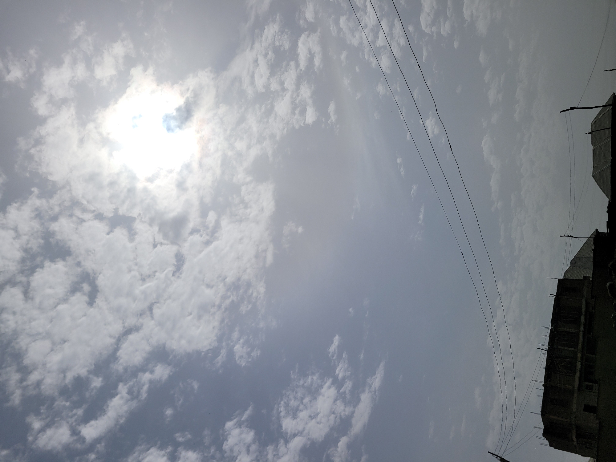 The image shows a daytime view of the sky in Barakin Akawo, Nigeria. Numerous small, white cumulus clouds cover most of the sky, with the sun shining brightly through a gap in the clouds on the left side of the frame. Several power lines stretch across the sky from the upper right to the lower left. The silhouette of a partially constructed building is visible in the lower right corner.