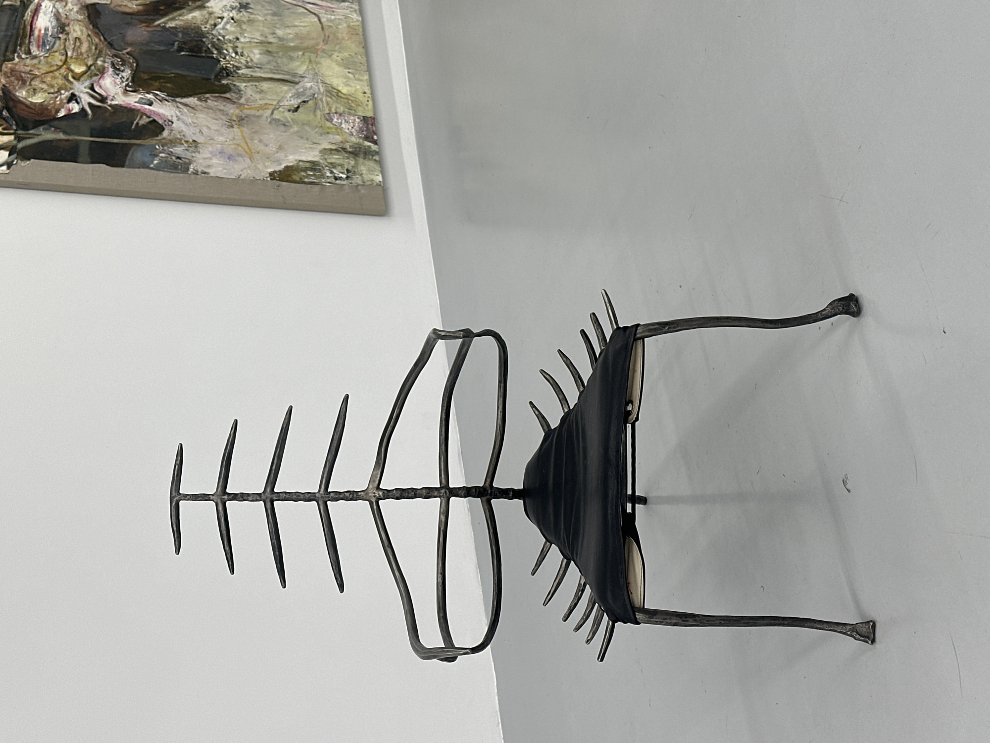 The image shows a metal sculpture resembling a fish skeleton placed on a white wall. An abstract painting hangs nearby on the upper left corner. The sculpture has a "spine" with ribs on one side and curved fins on the other. In the center is what looks like a black folded fabric or leather item. The background is a plain white wall, suggesting an indoor location. There are no people visible. The location is the city of London, United Kingdom.