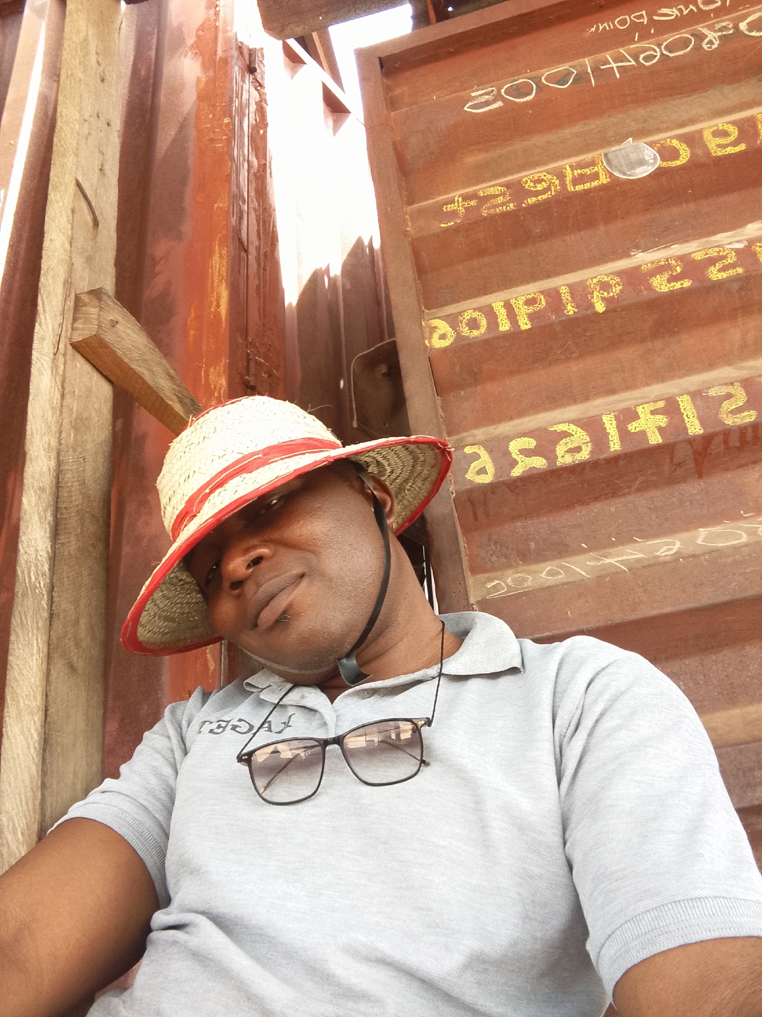 A man with dark skin and a straw hat is in an outdoor environment, possibly near shipping containers or metal structures. He is wearing a grey polo shirt and has glasses hanging on his shirt. The background shows weathered metal panels with some writing or markings. The location in the city of Unknown, Unknown cannot be determined.