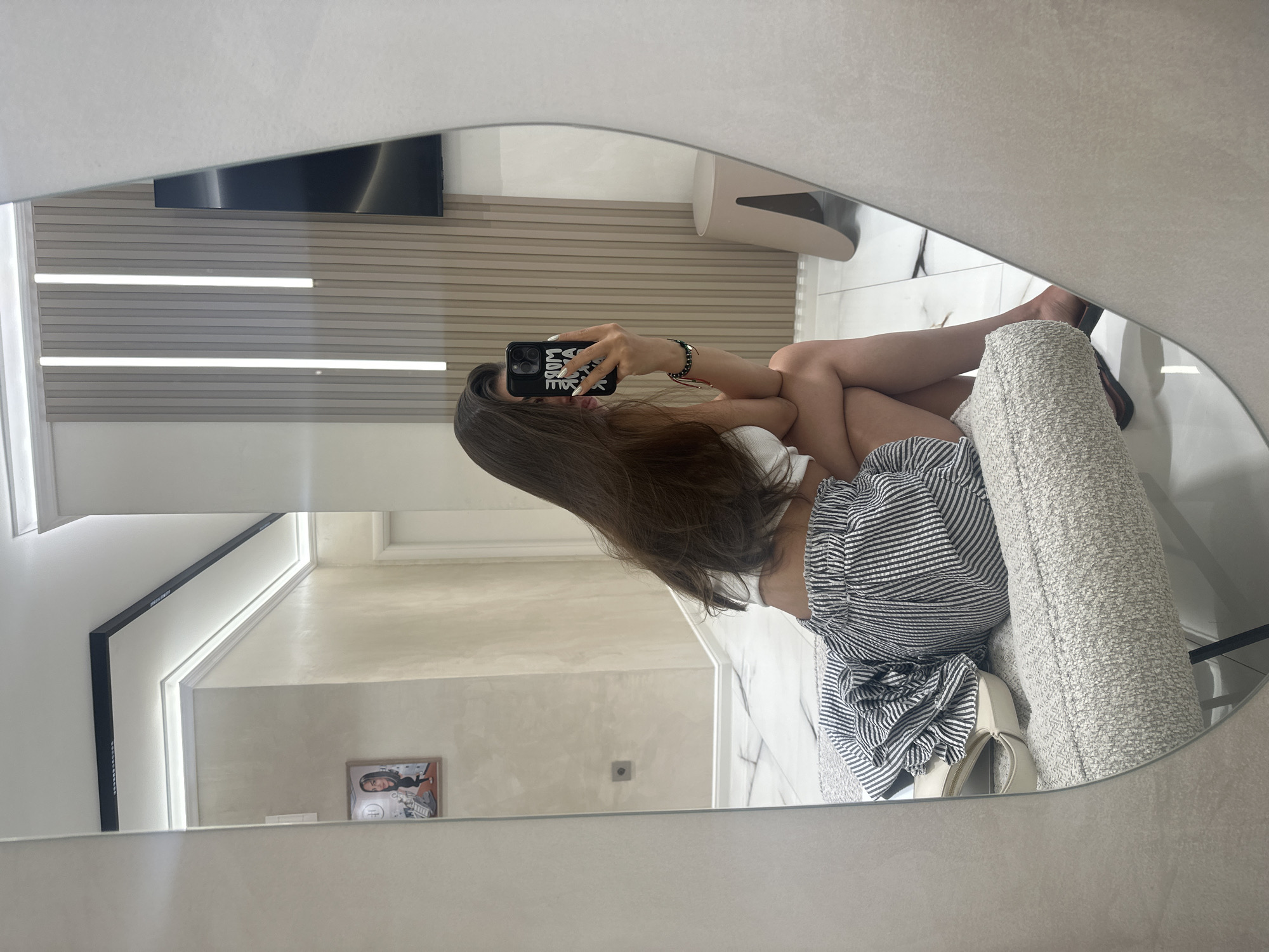 A woman is taking a selfie in a mirror in a room with marble floors and a textured wall. She is wearing a white crop top and striped shorts, and her hair is long and brown. A pair of cream-colored sandals and a striped garment are on a bench next to her. The room also has a framed picture on the wall and a modern stool. The mirror is large and oval-shaped. The scene is captured in a brightly lit interior space that appears to be a fitting room or a well-decorated home.