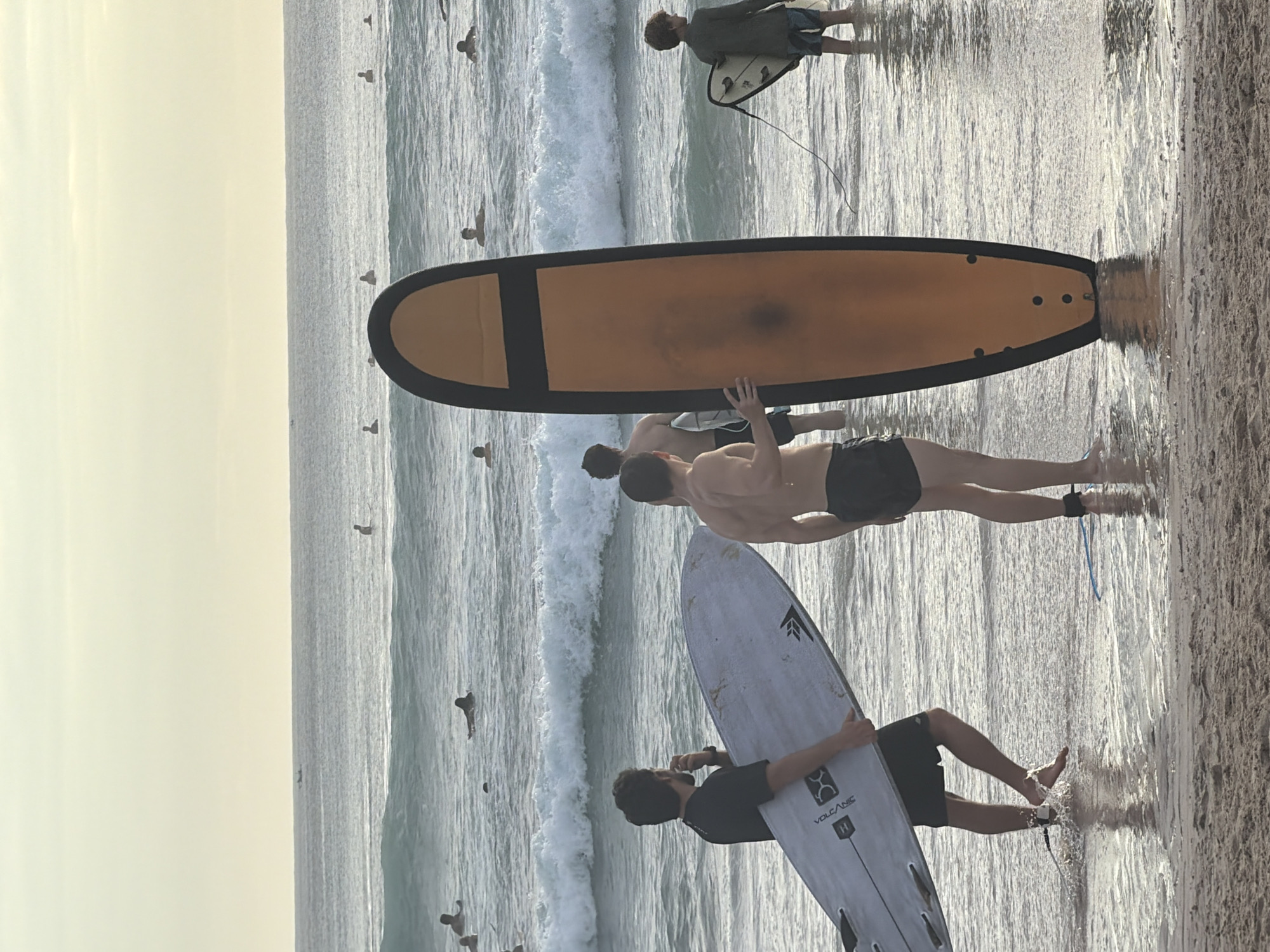 The image shows a beach scene in Kuta Selatan, Indonesia, with several people and surfboards. Four people are visible, three are standing in the shallow water near the shore, holding surfboards, and one is further out in the water. One person in the foreground is holding a white surfboard with a black logo, and is wearing a black shirt and dark shorts. Behind this person are two other people. One is shirtless, wearing black shorts, standing next to a large orange surfboard. In the water further out is a person with a smaller board. The ocean is wavy, and the sky is overcast. A flock of seabirds is visible in the sky above the ocean.