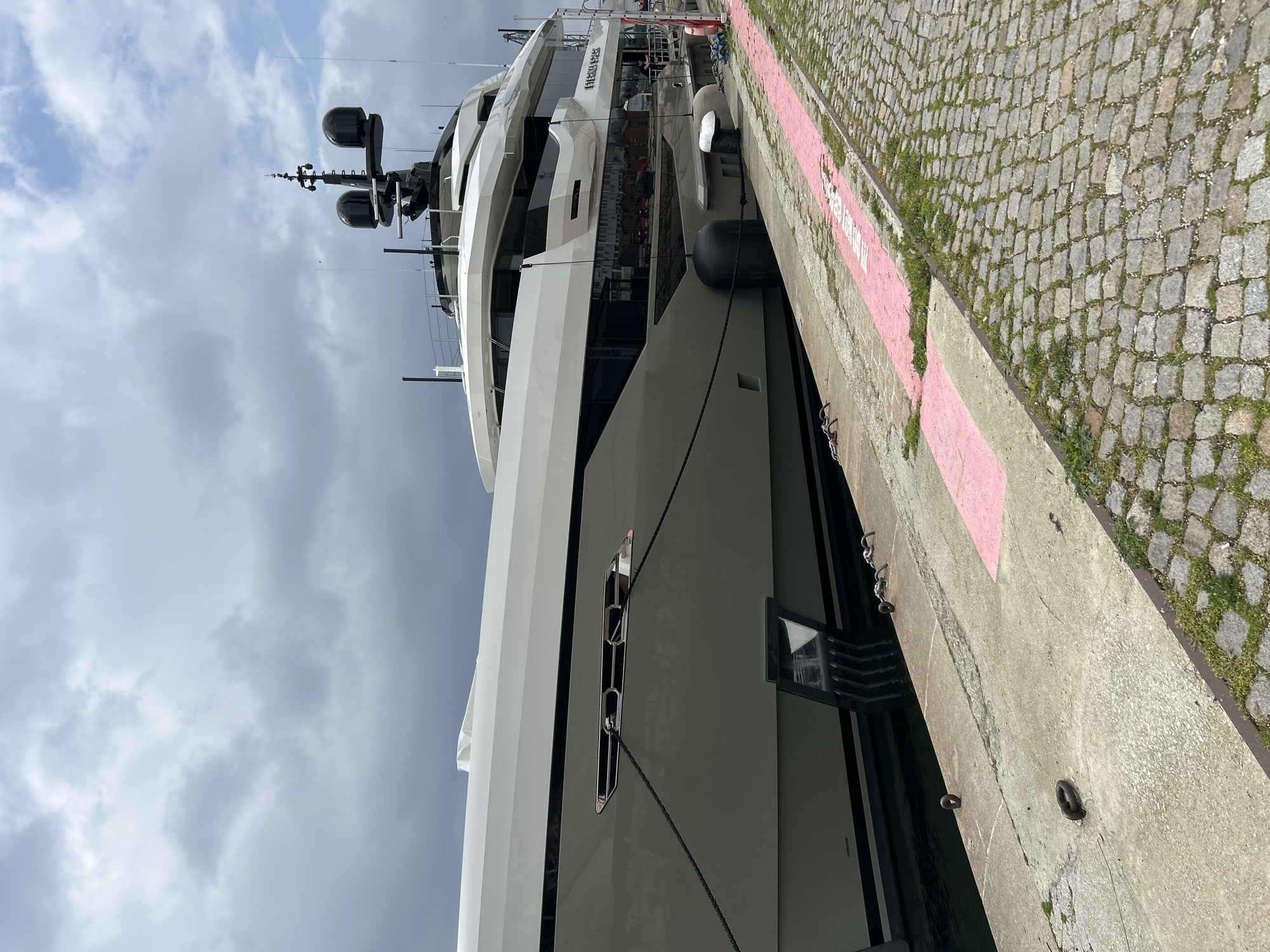 A large yacht is docked alongside a concrete pier. The yacht is white and gray, with multiple decks and antennas. It is secured to the pier with thick black ropes. The pier has a pink stripe painted along the edge, and is lined with cobblestones. The sky is cloudy. The city is Unknown.